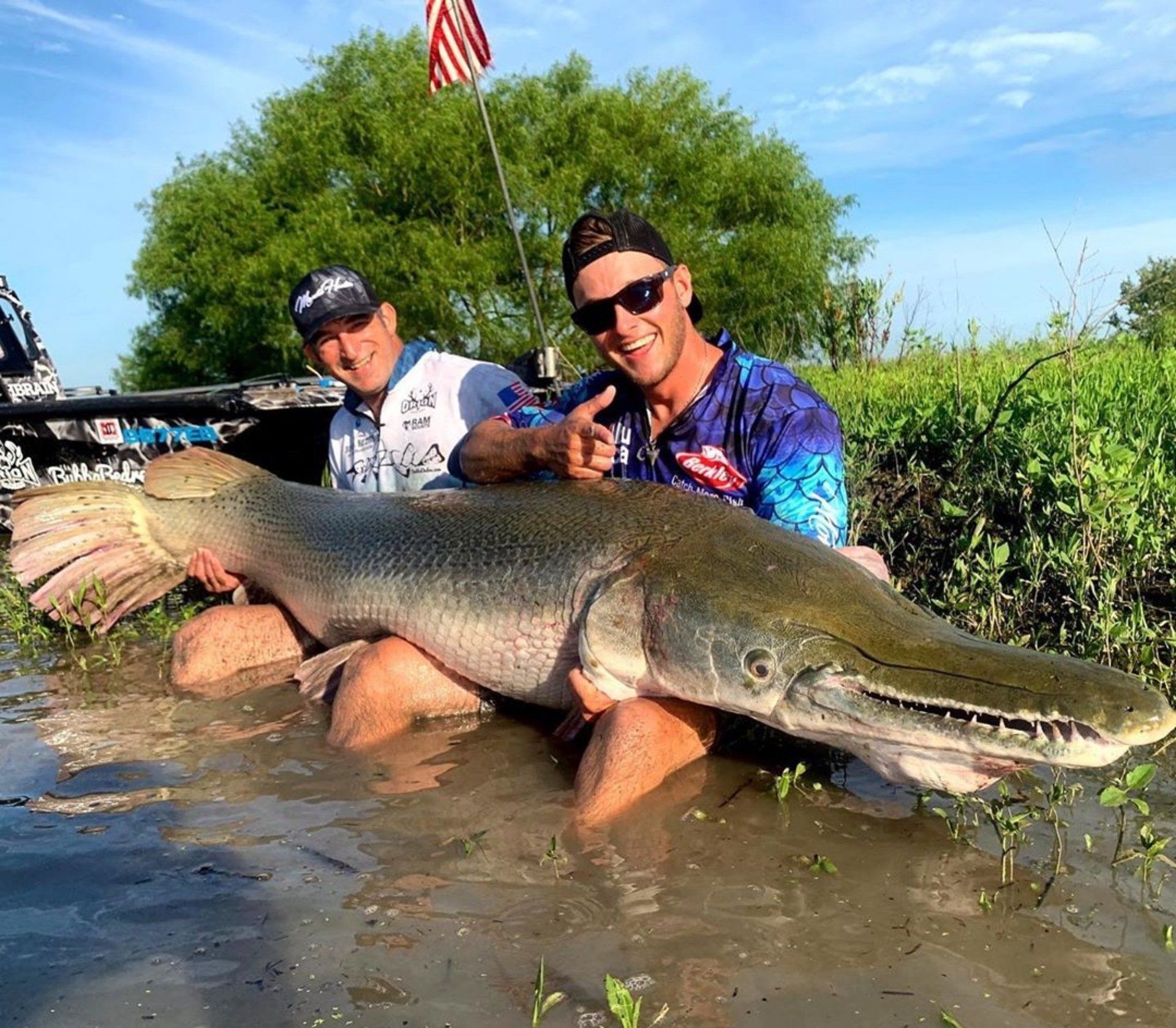 Two men in water holding a giant gar fish, smiling. Green foliage and an American flag in the background.