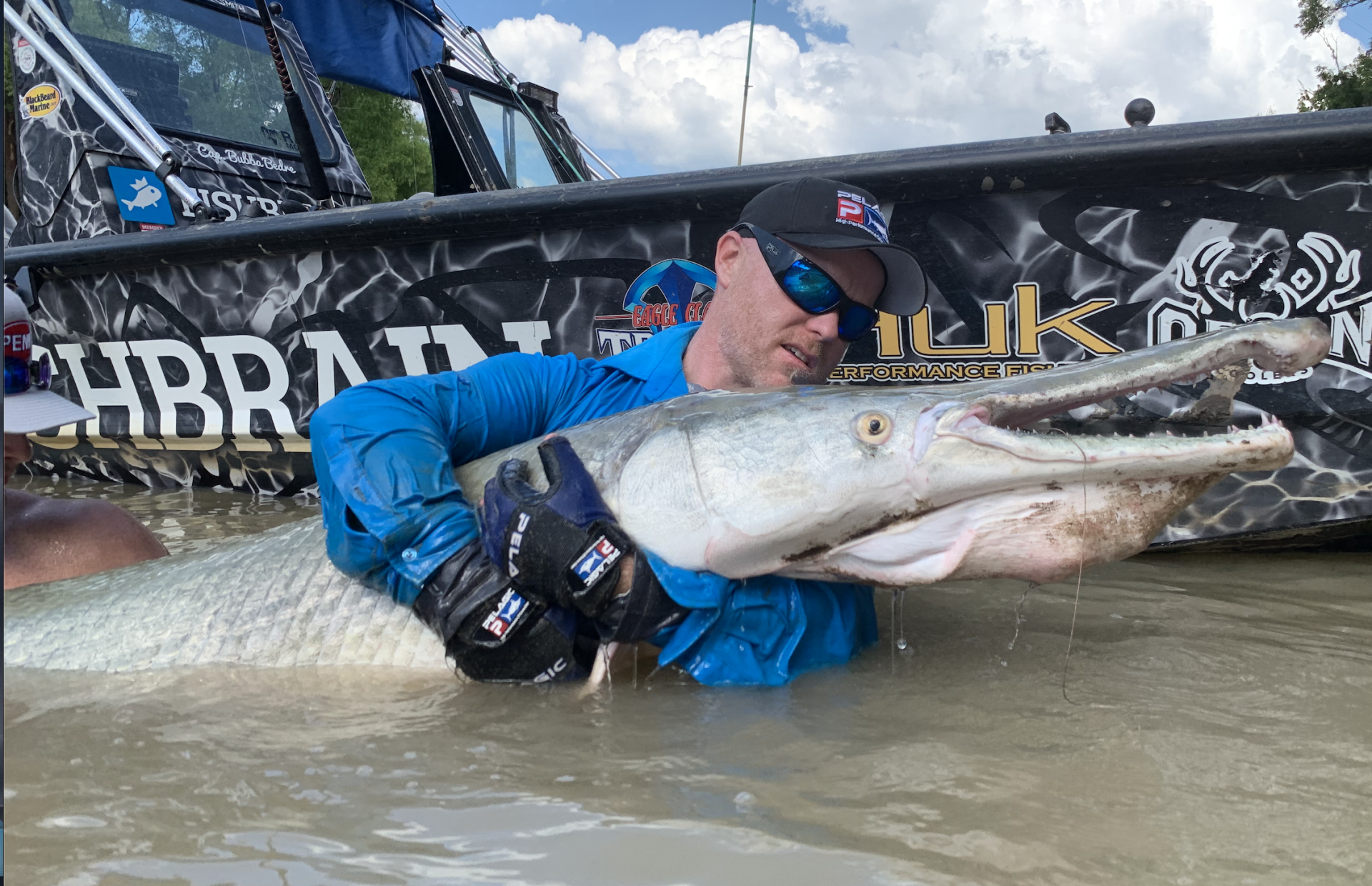 Man holding a large alligator gar in water, wearing a blue shirt and sunglasses. Boat in the background.