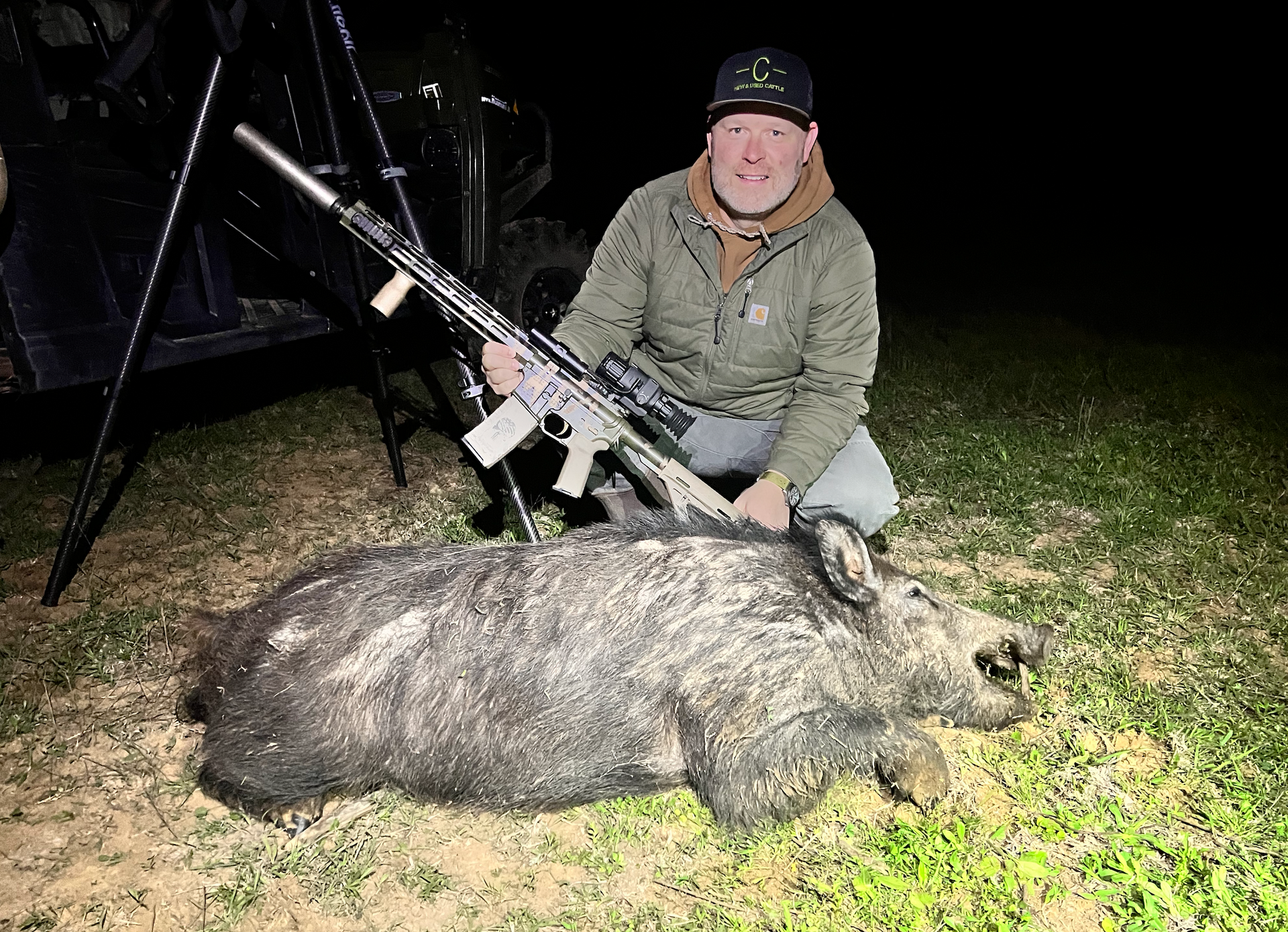 Man kneels with rifle next to a harvested wild hog in a grassy setting at night.