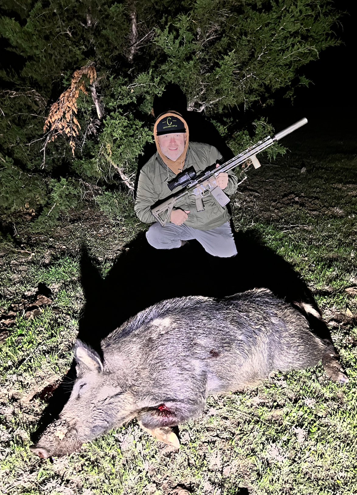 Man kneeling with a rifle next to a large, dead wild pig at night.
