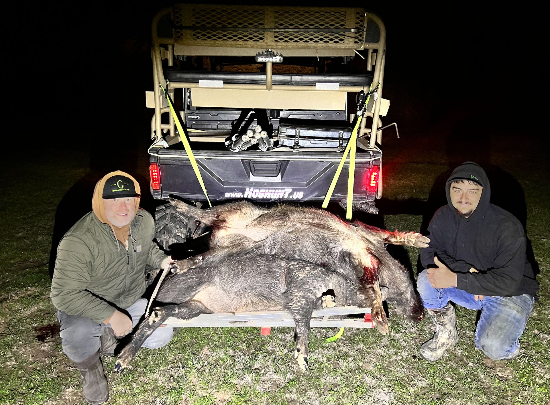 Two men kneel with a utility vehicle behind them and several dead wild hogs. Nighttime.