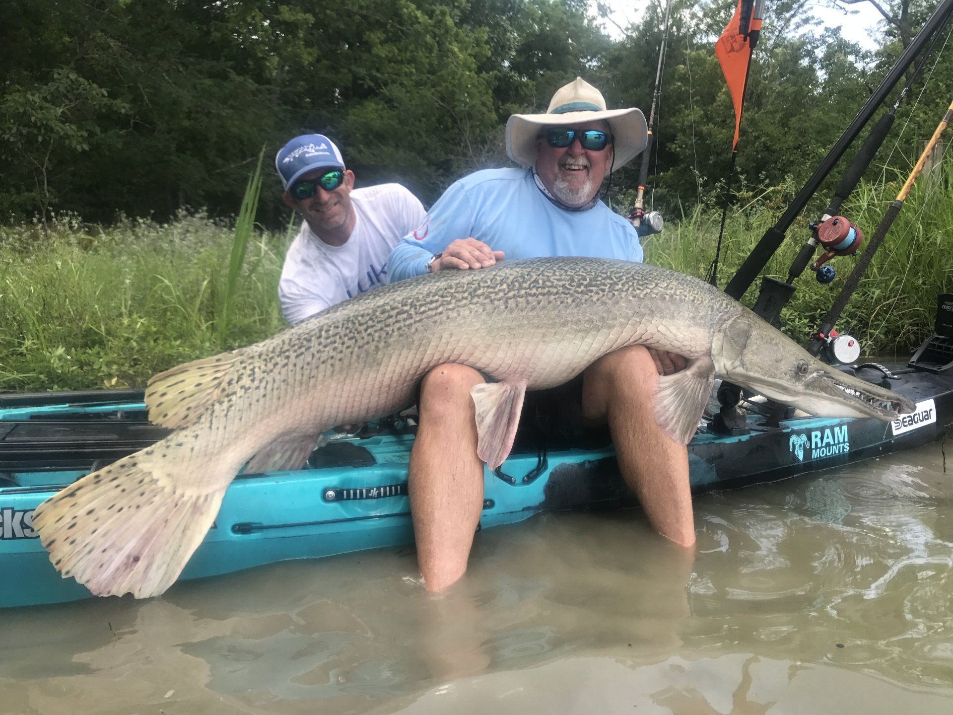 Two men on a kayak holding a large alligator gar fish. Men are smiling in a green river.