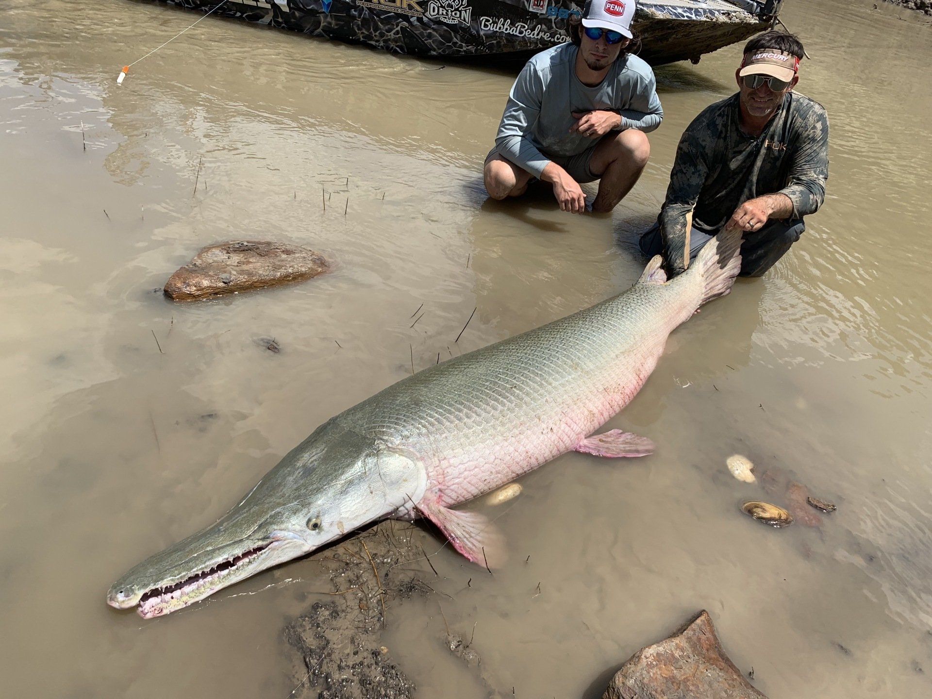 Two men pose with a large alligator gar fish on a muddy bank near a boat.