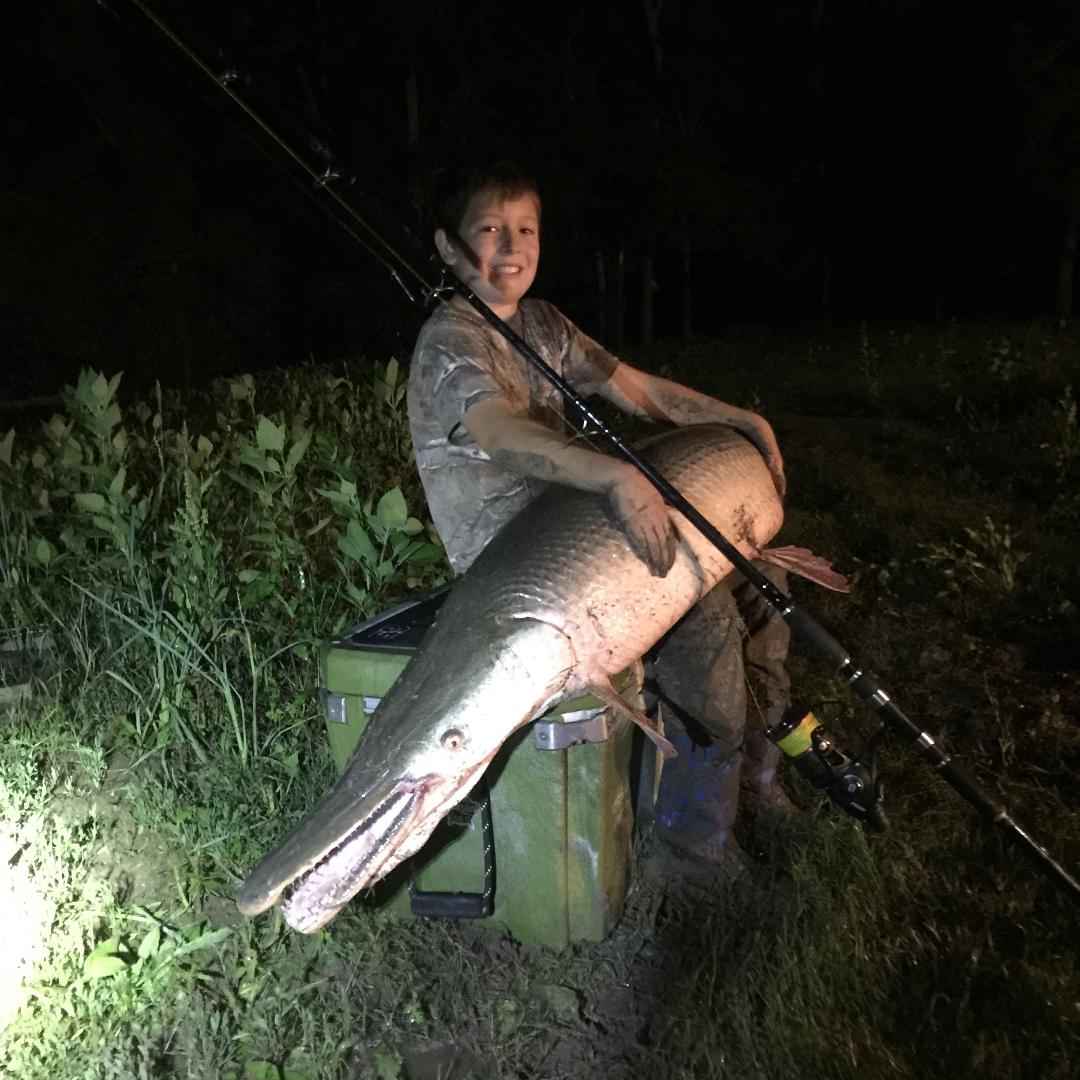 Boy holds a large alligator gar fish at night, smiling near a fishing rod and gear.