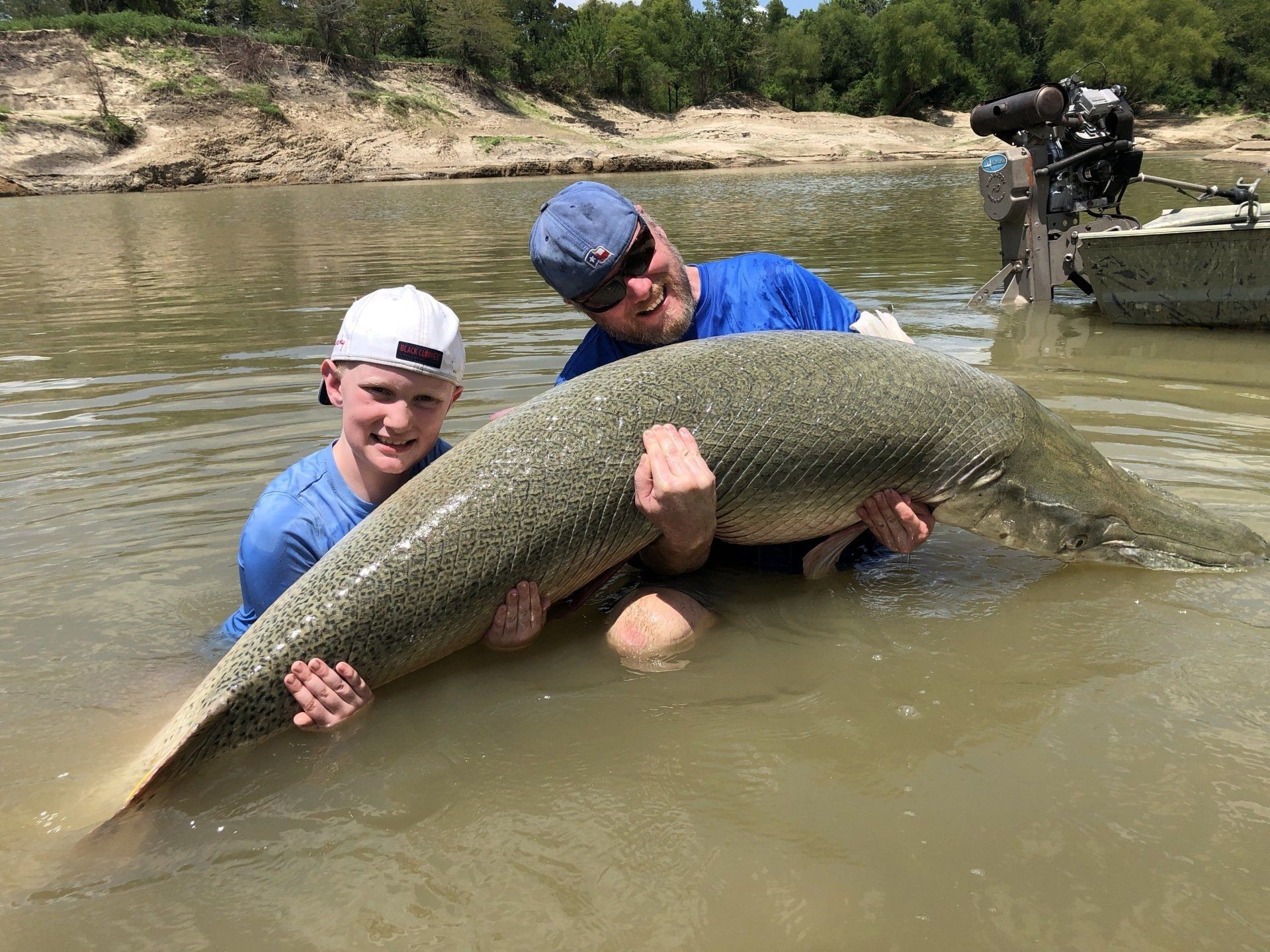 Two men holding a large, spotted fish in shallow water; boat in the background.