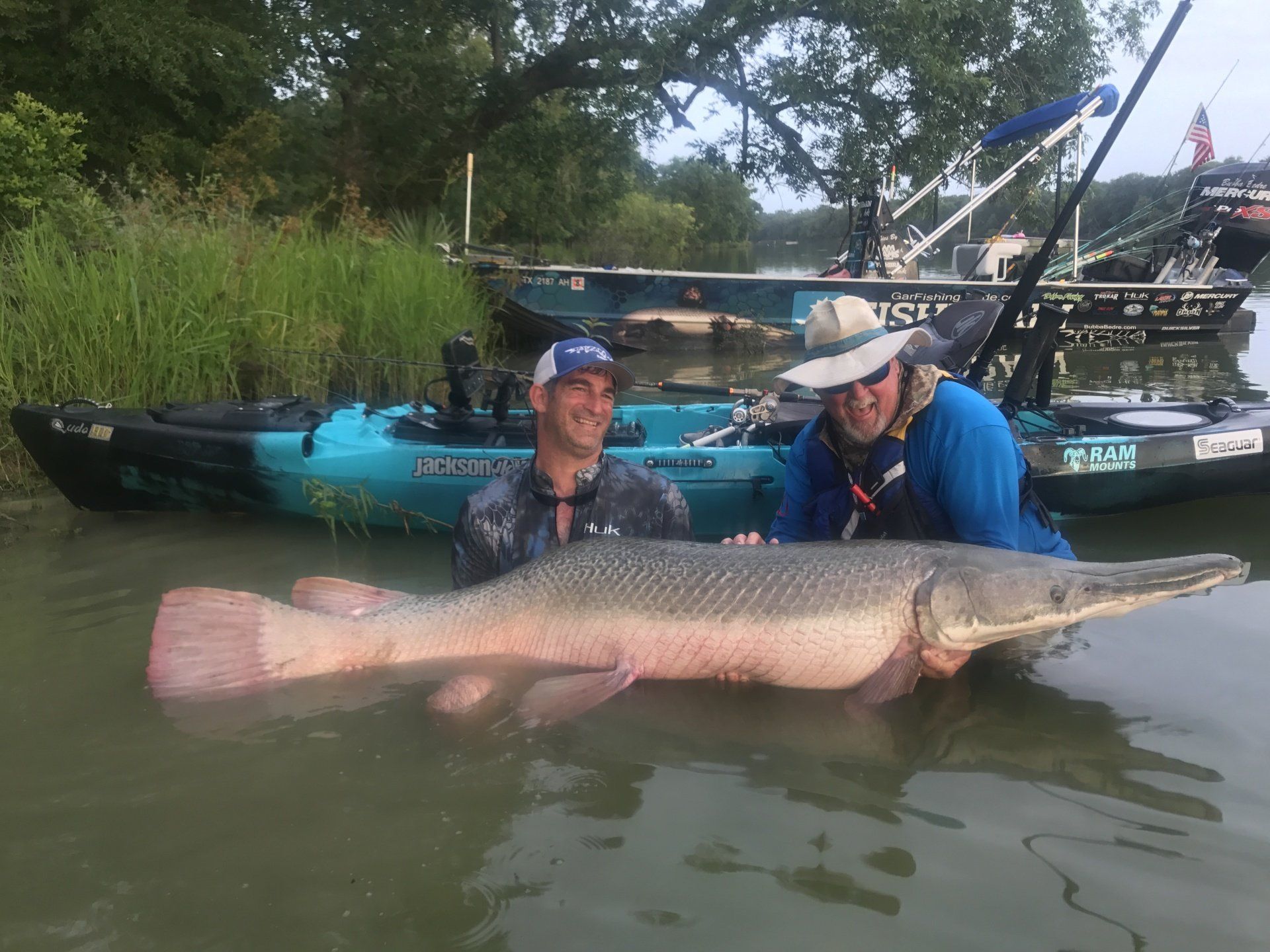 Two men in water holding a large alligator gar fish near kayaks on a riverbank.