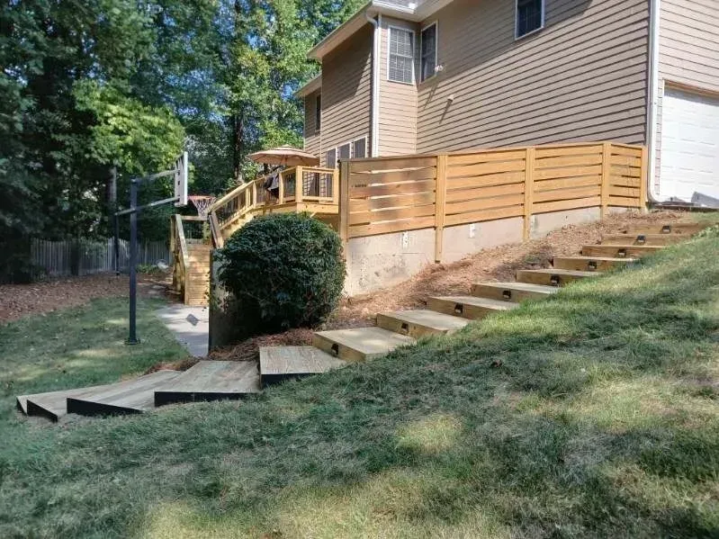 Wooden steps leading up a grassy hill to a deck with horizontal wooden fencing and a house.
