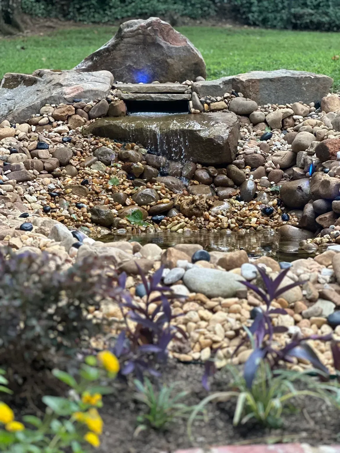 Water cascading over rocks into a small pond, surrounded by pebbles and purple plants in a garden setting.