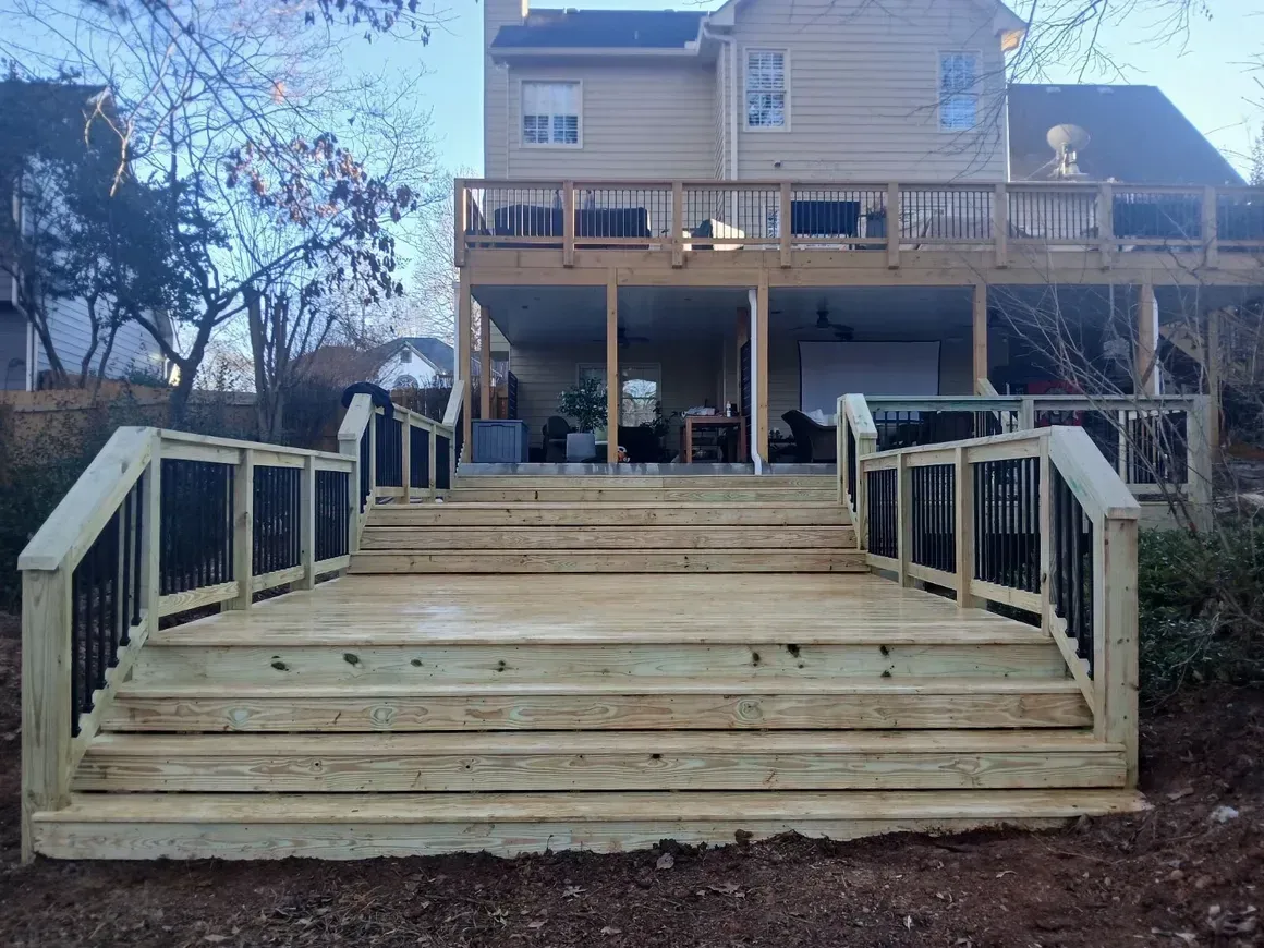 Wooden deck with steps leading to a two-story house. Black railings and bare trees surround.