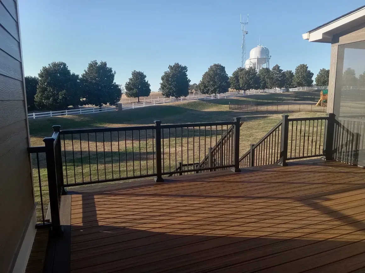 A wooden deck with black railing overlooking a field, trees, and a white building under a blue sky.