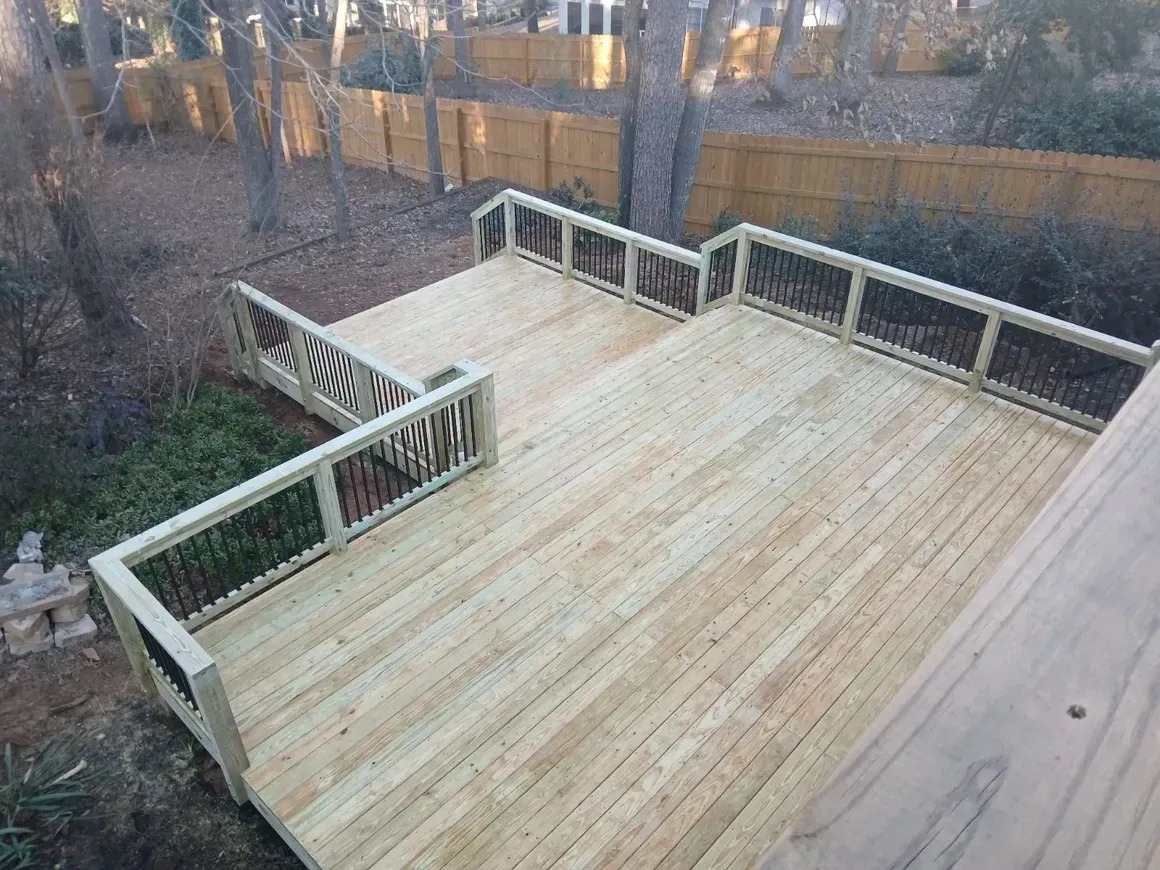 Wooden deck with railings, built at different levels, in a backyard setting, surrounded by trees and a wooden fence.