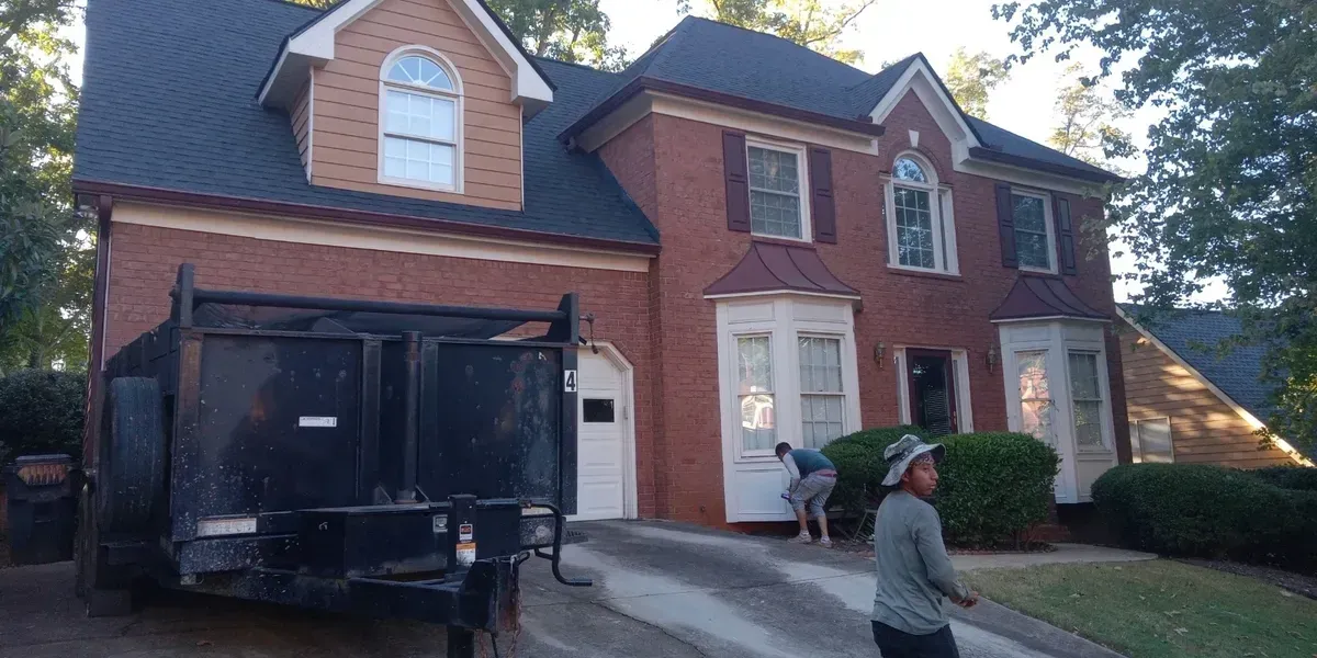 A brick house with a dark roof and a dumpster in the driveway. Two people are working near the entrance.