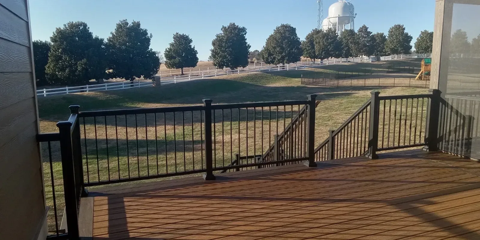 View from a deck with black railing overlooking a green lawn, trees, and a white water tower.