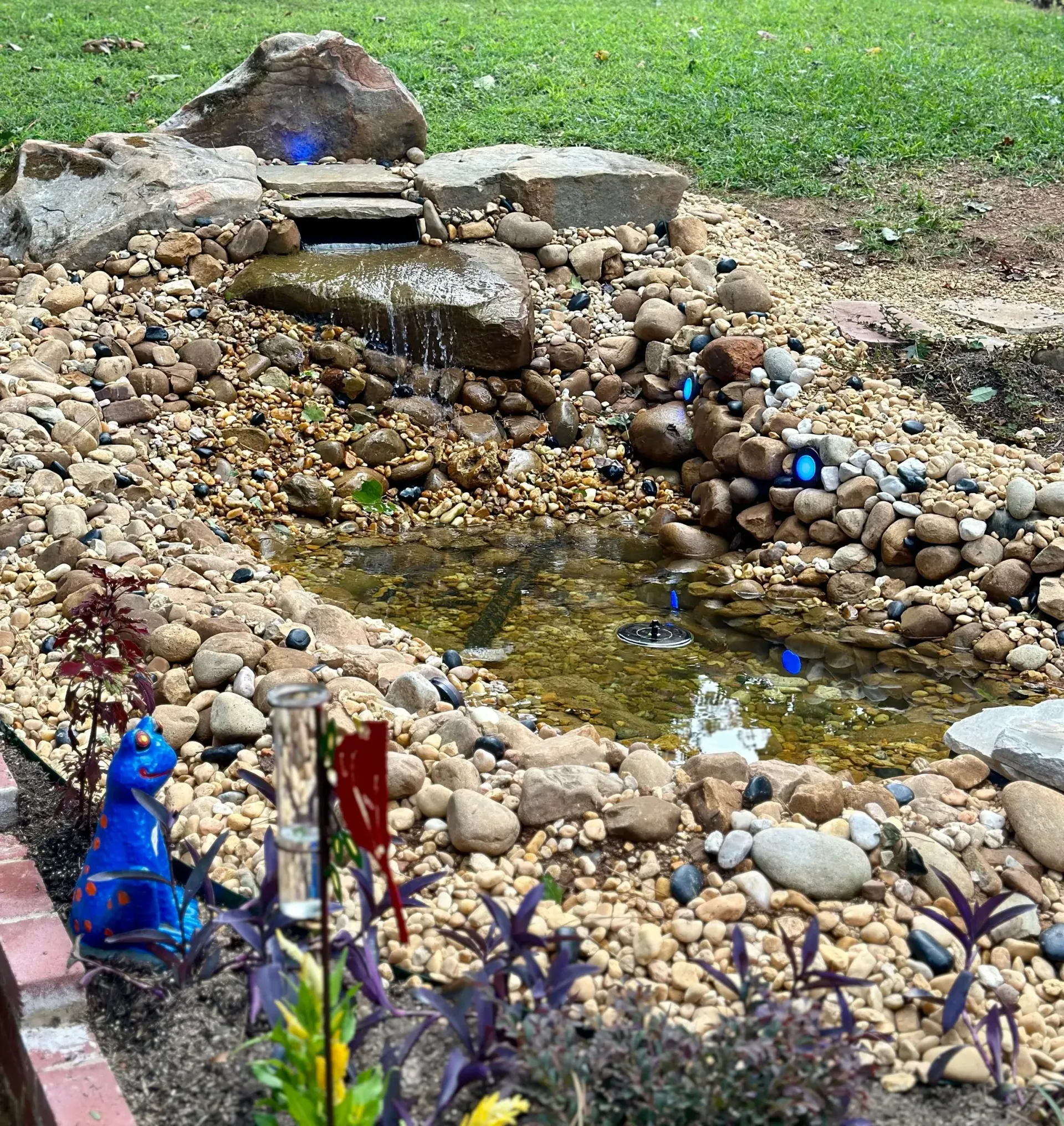 Water feature with rocks, small waterfall, and a blue cat statue.