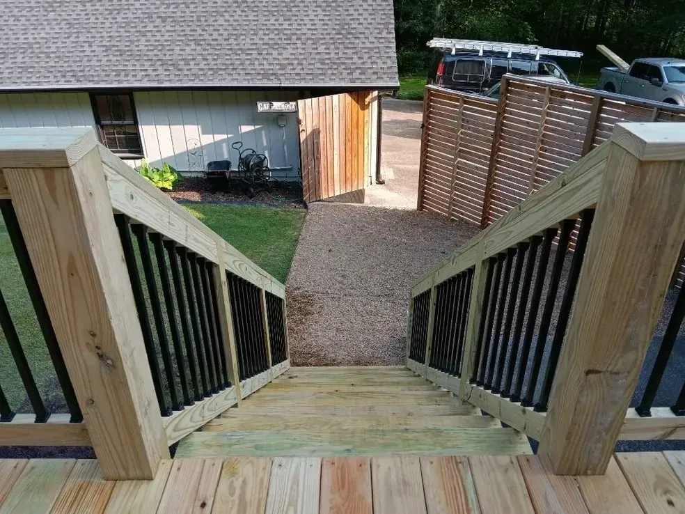 Wooden deck with stairs leading down to a gravel path, flanked by black railings and a wooden fence.