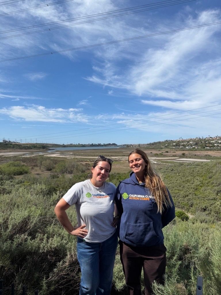 Four people pose with tools and a truck near a hillside. Cloudy sky, dirt ground.