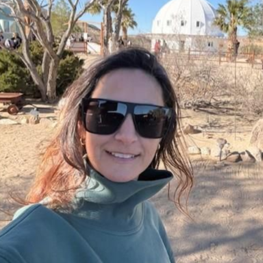 Woman with long brown hair, smiling, wearing a white t-shirt, outside with a blurred background.