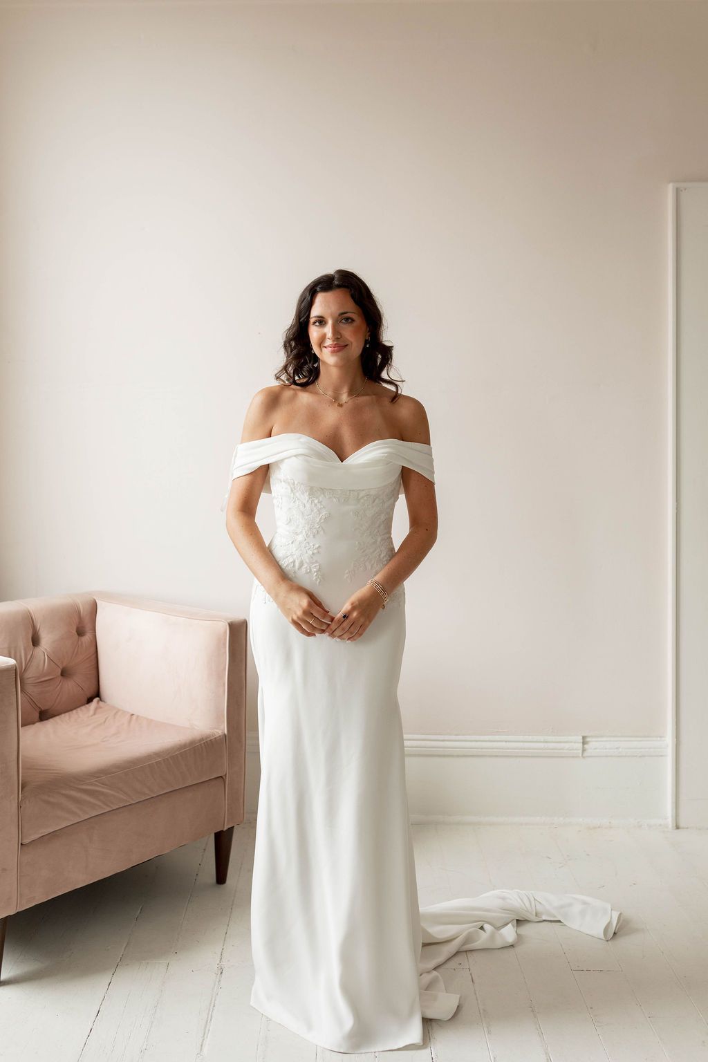 Bride in a white off-shoulder gown standing in a bright room beside a blush pink sofa