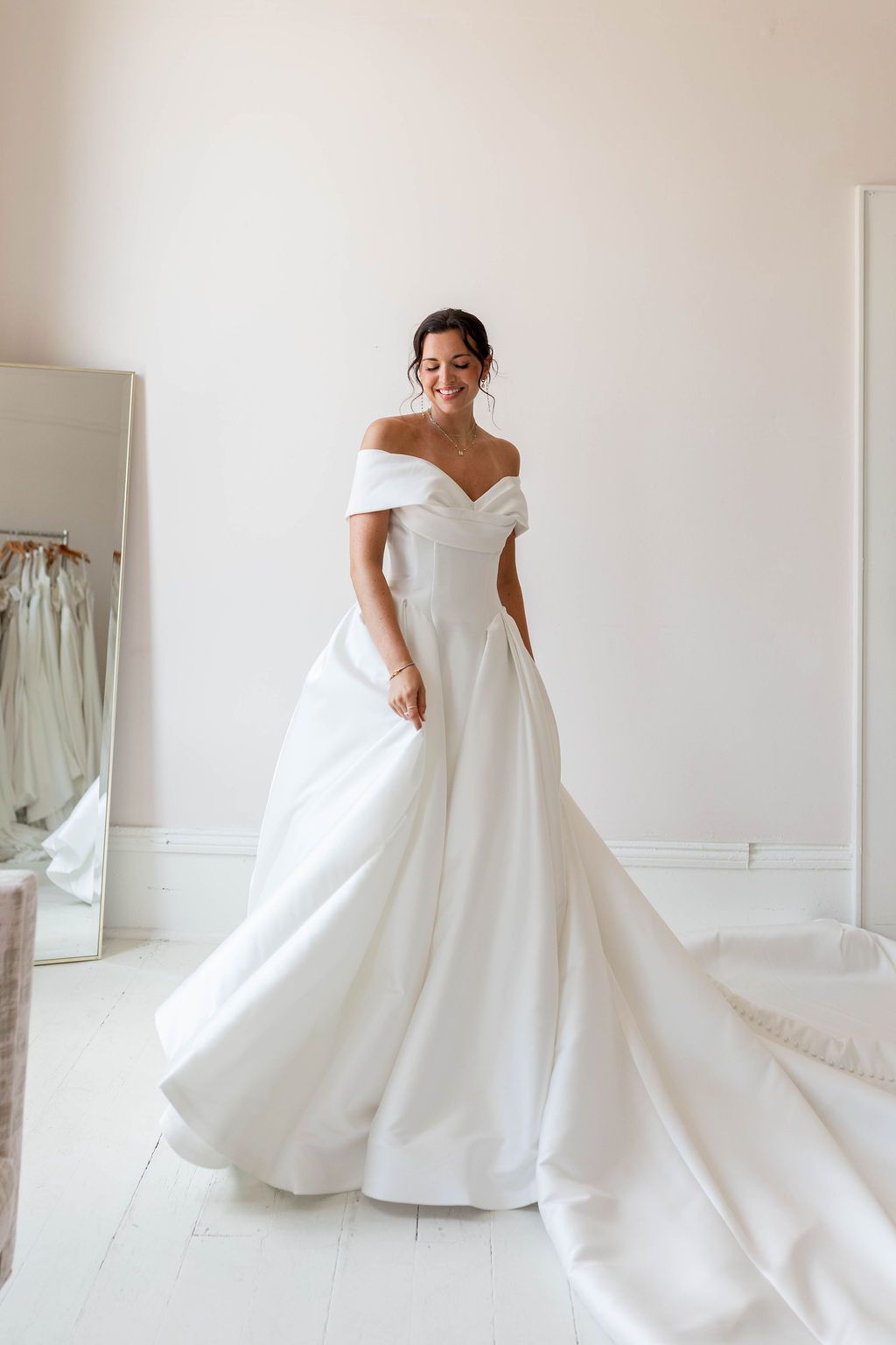 Bride in an off-shoulder white wedding gown standing in a bright room with a long flowing train