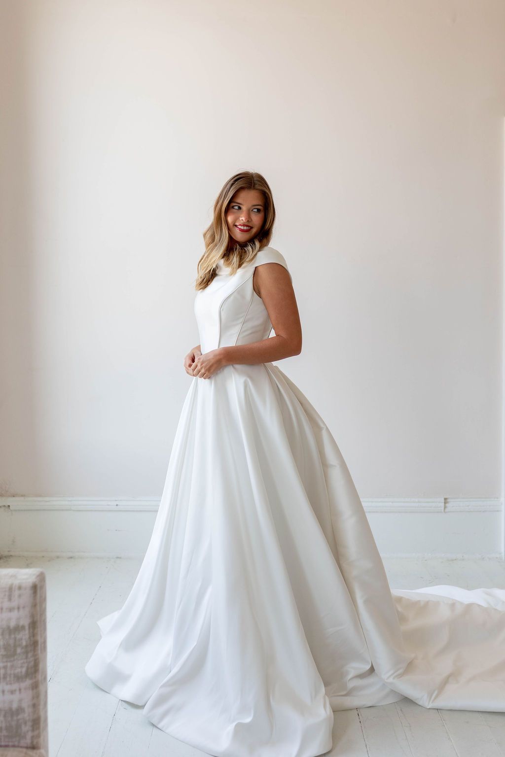 Woman in a white wedding gown standing in a bright, minimalist room, smiling gently.
