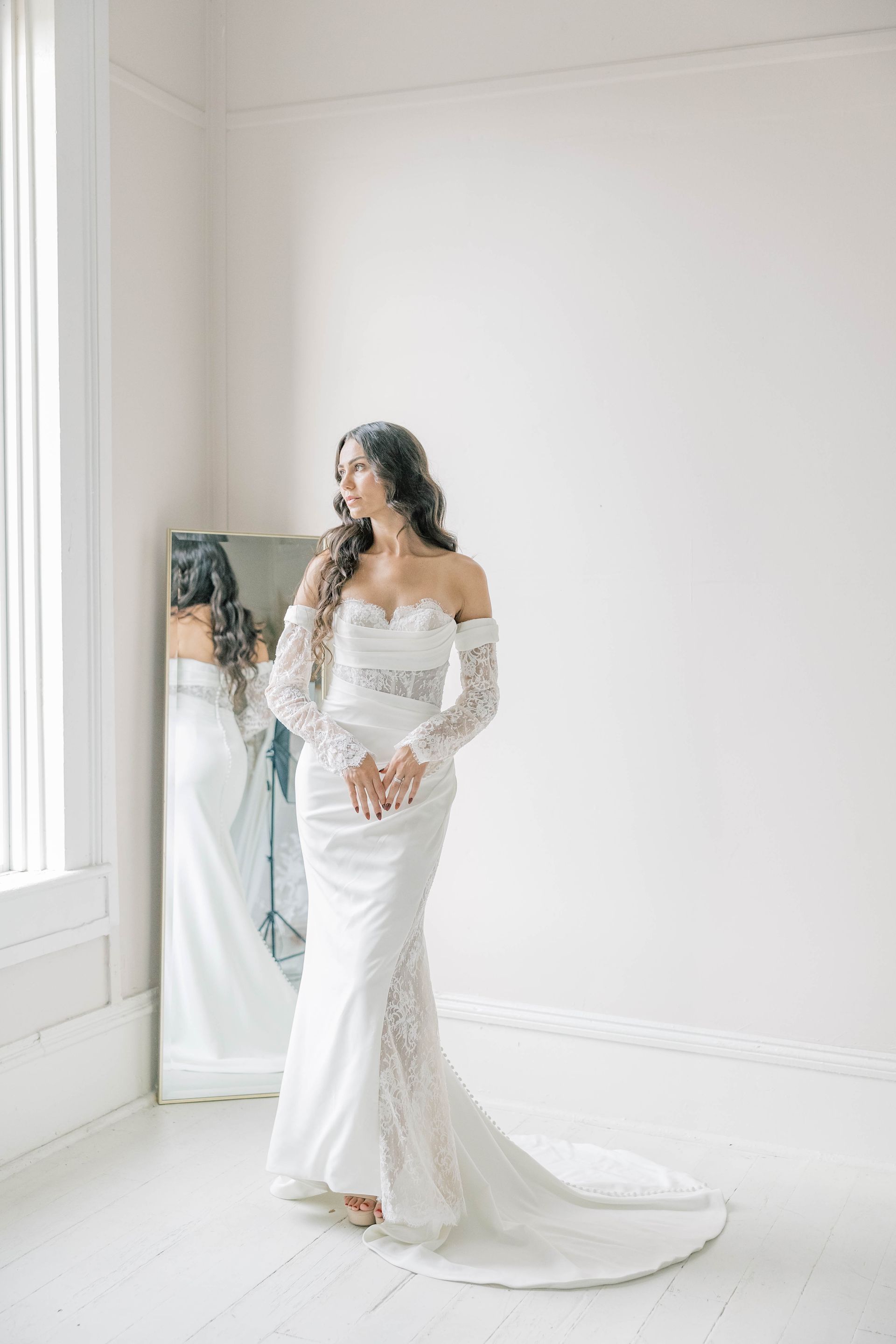 Bride in white lace wedding dress looks towards window, standing by mirror.