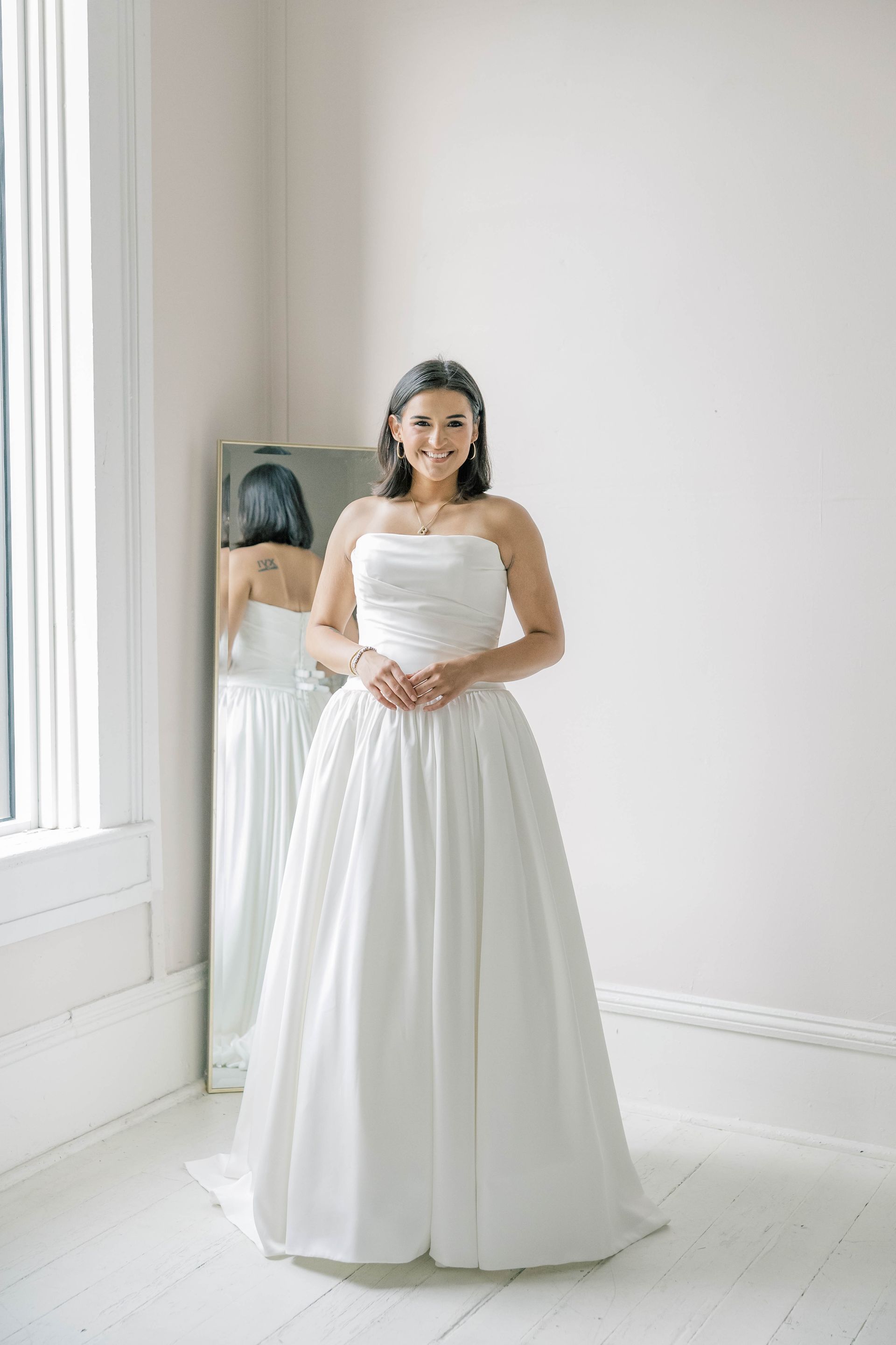 Woman in strapless white wedding dress, smiling, in front of a mirror. Light-filled room.
