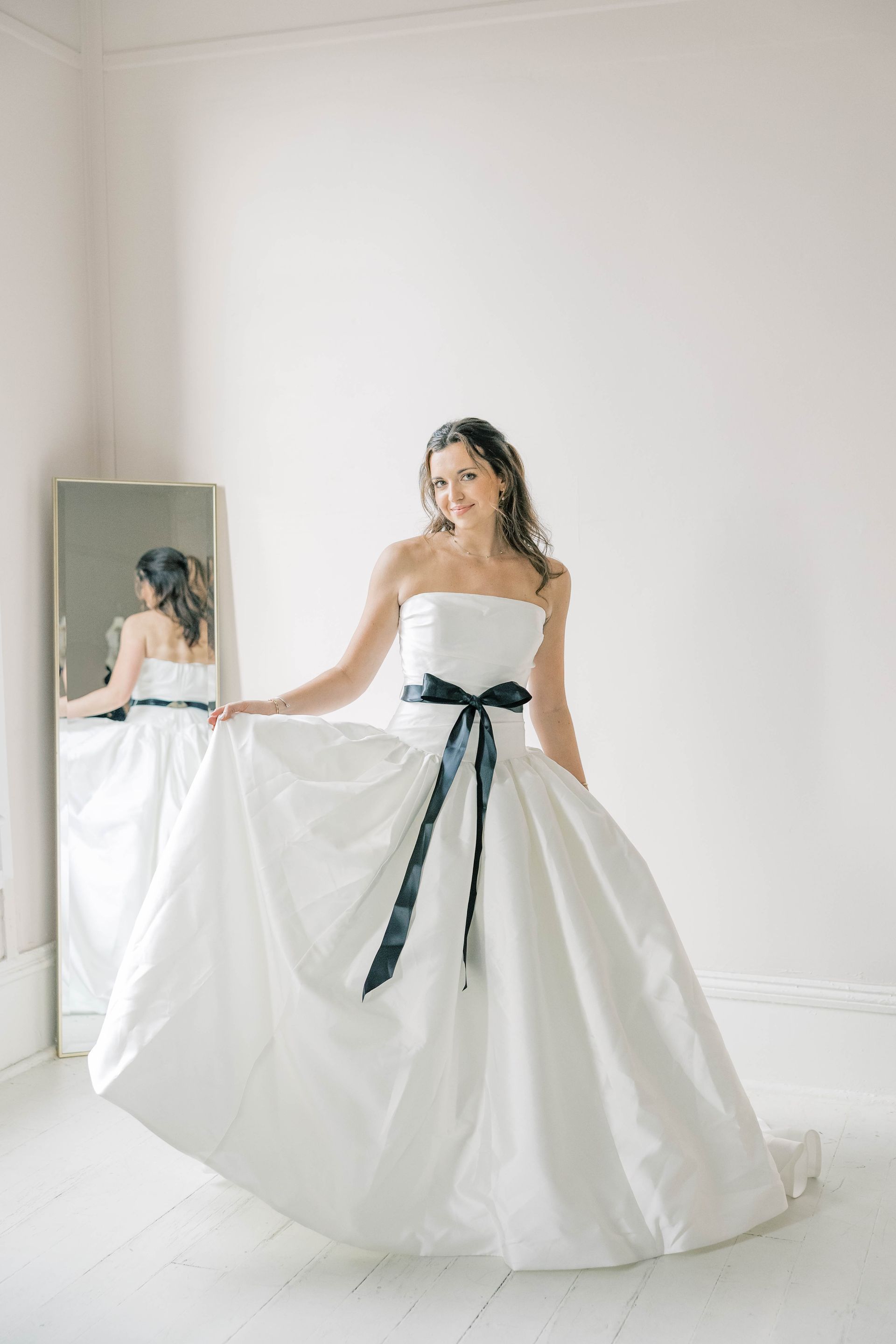 Woman in white strapless gown with black sash, posing in front of a mirror. Light, airy room.