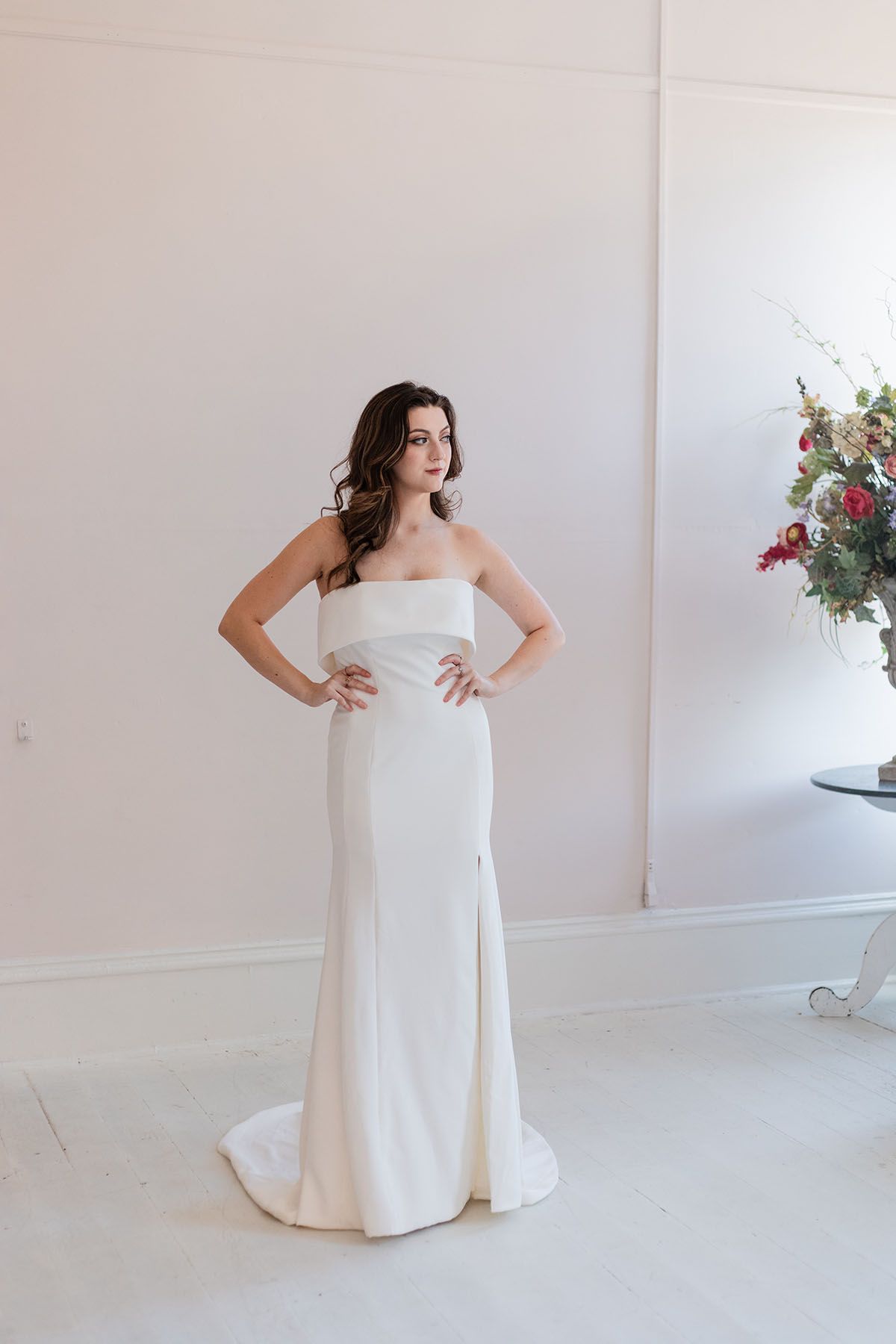 A woman in a white wedding dress is standing in a room with her hands on her hips.