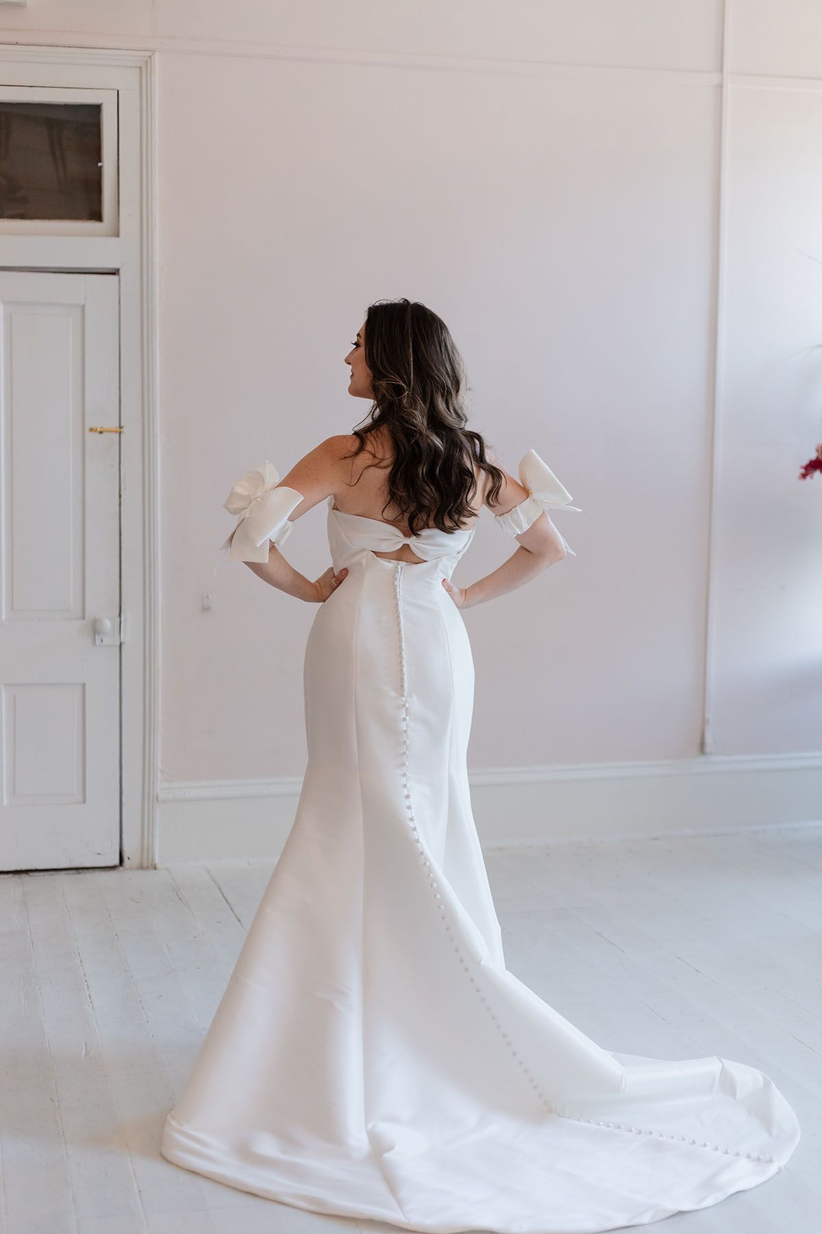 A woman in a white wedding dress is standing in a room with her hands on her hips.