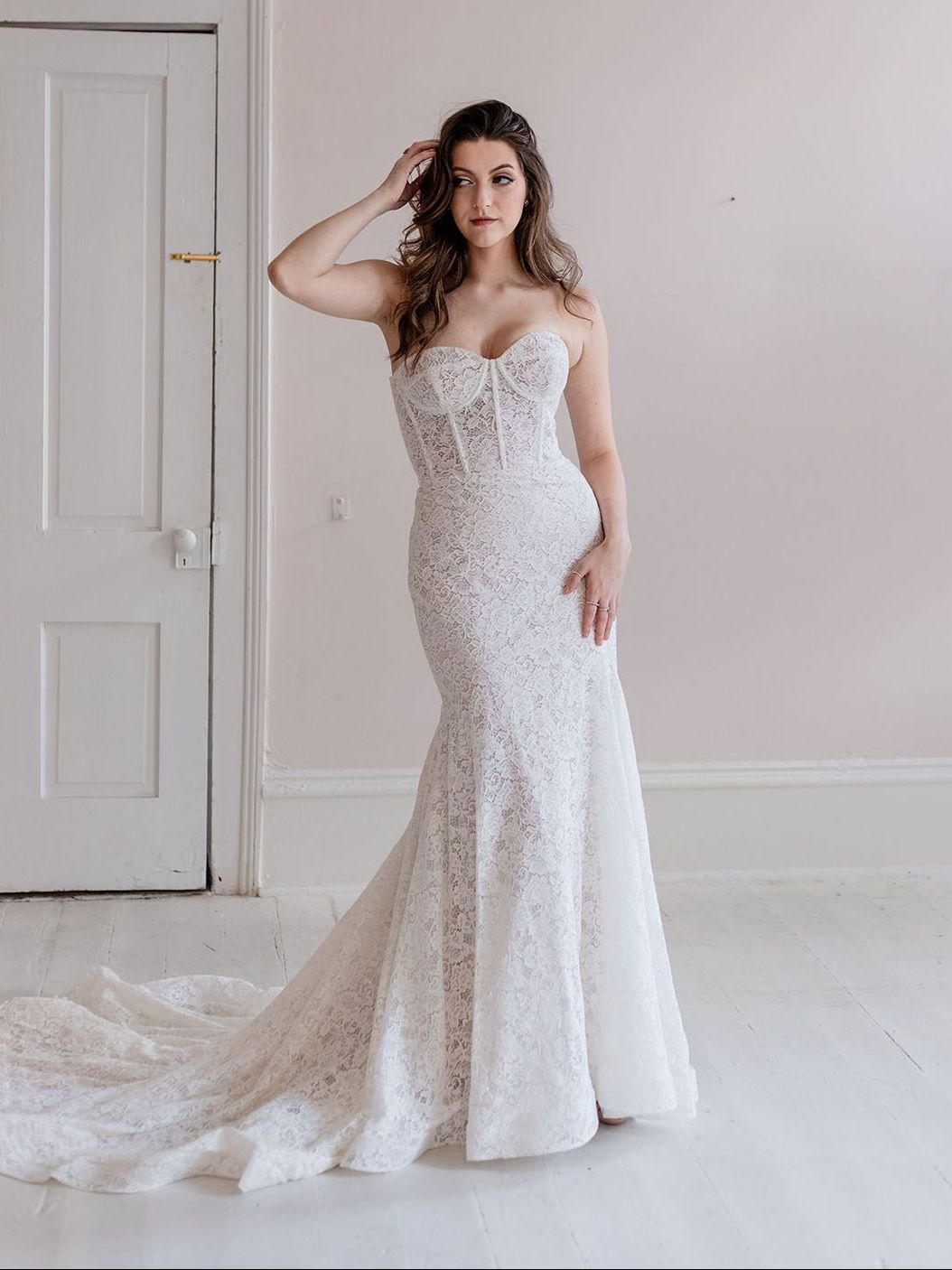 A woman in a white lace wedding dress is standing in front of a door.