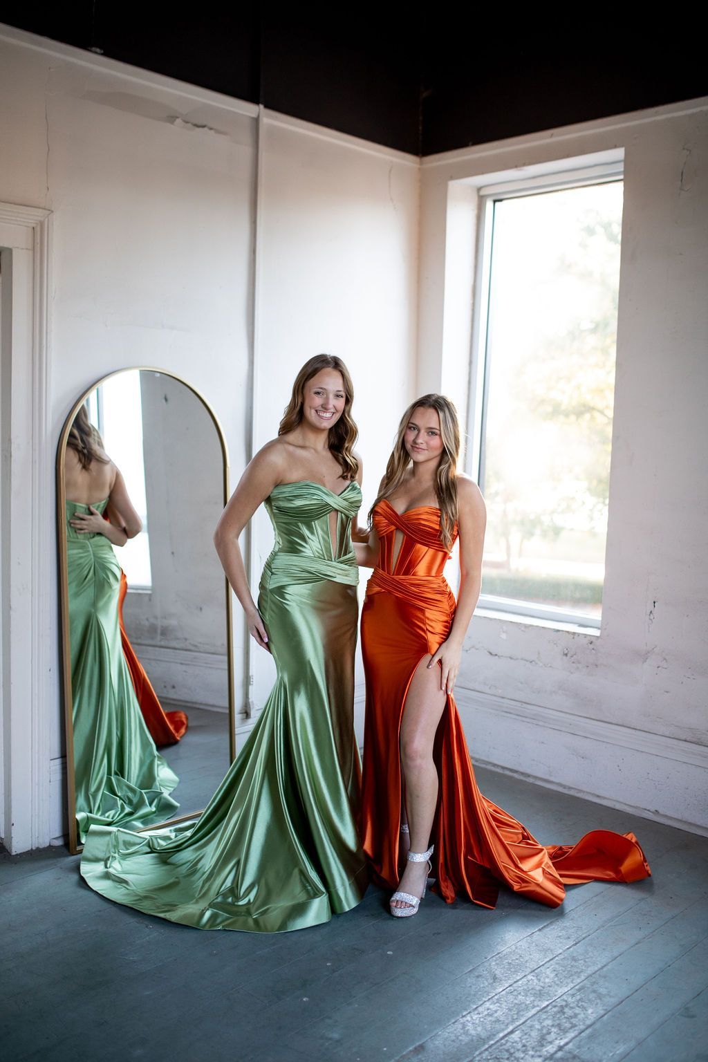 Two women in long dresses are standing next to each other in front of a mirror.