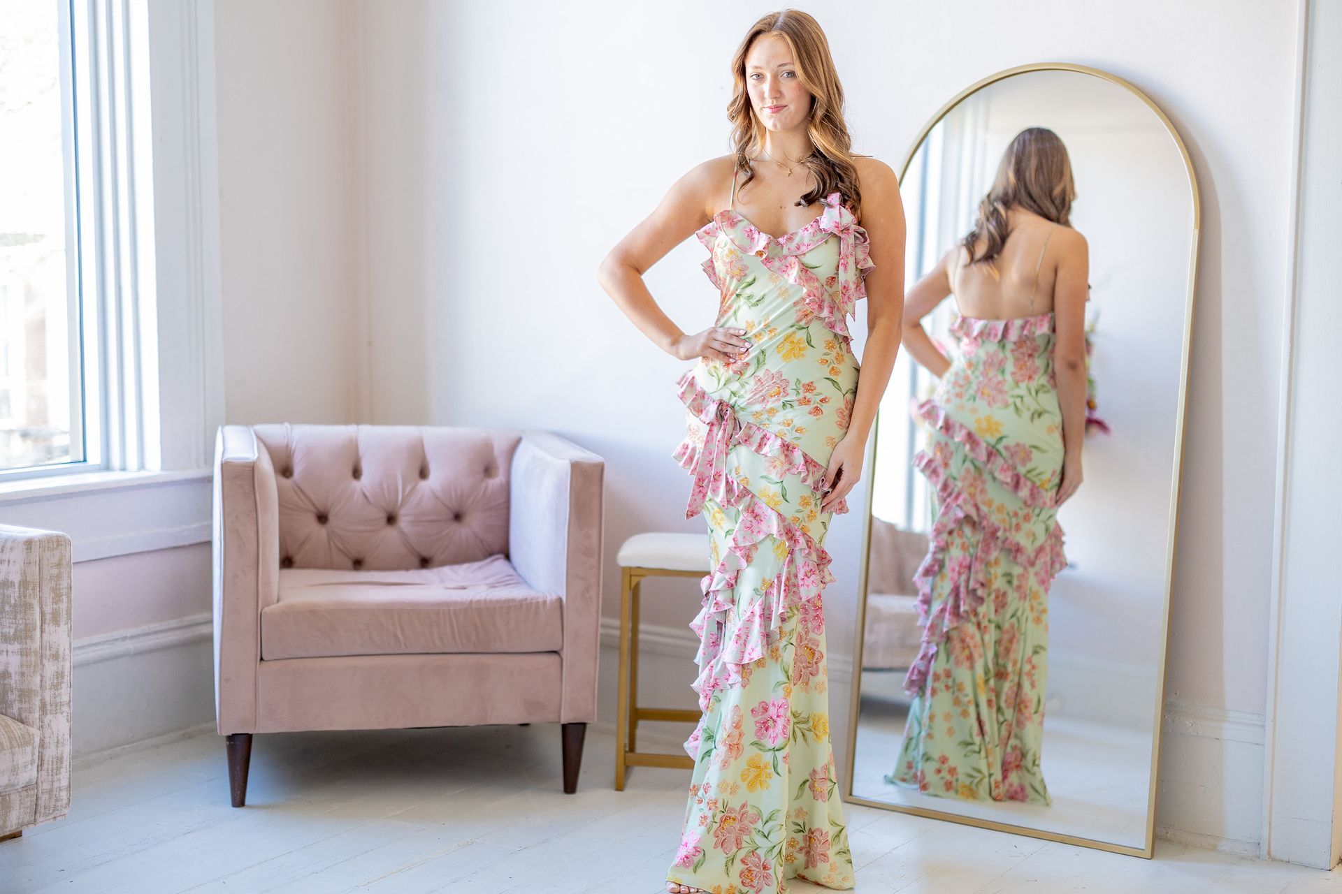 Woman in floral gown poses by a mirror in a light-filled room with pink accents.