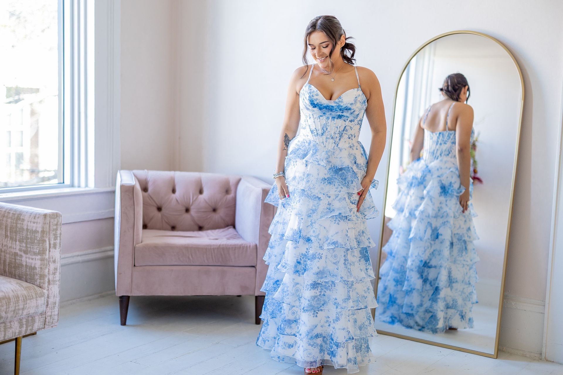 Woman in blue and white floral dress looking at herself in a gold-framed mirror, next to a pink chair.