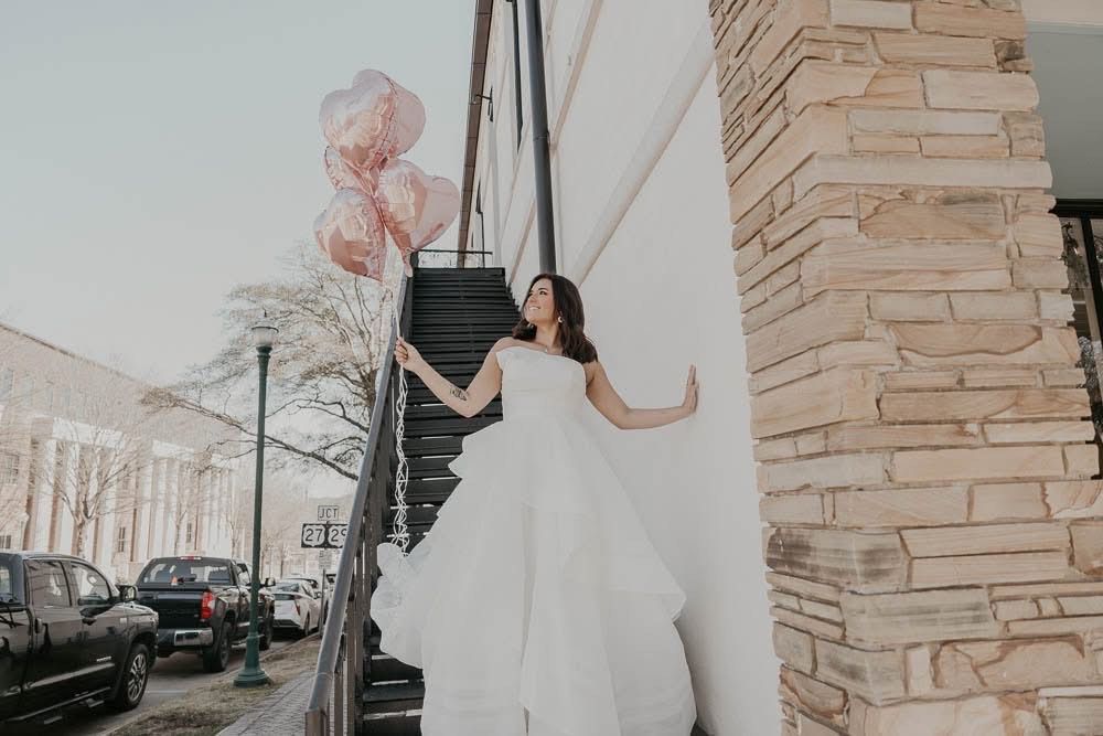 Woman in wedding dress holding heart balloons on outdoor staircase.
