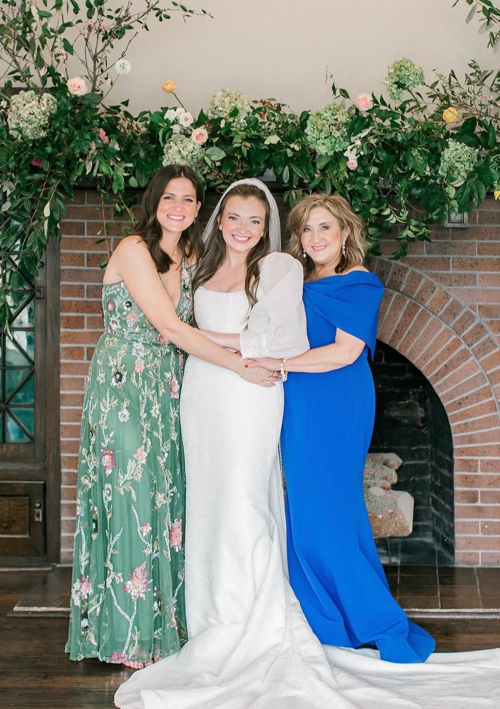 Bride with mother and sister in front of a fireplace, floral arch. Woman in white dress flanked by women in blue and green.