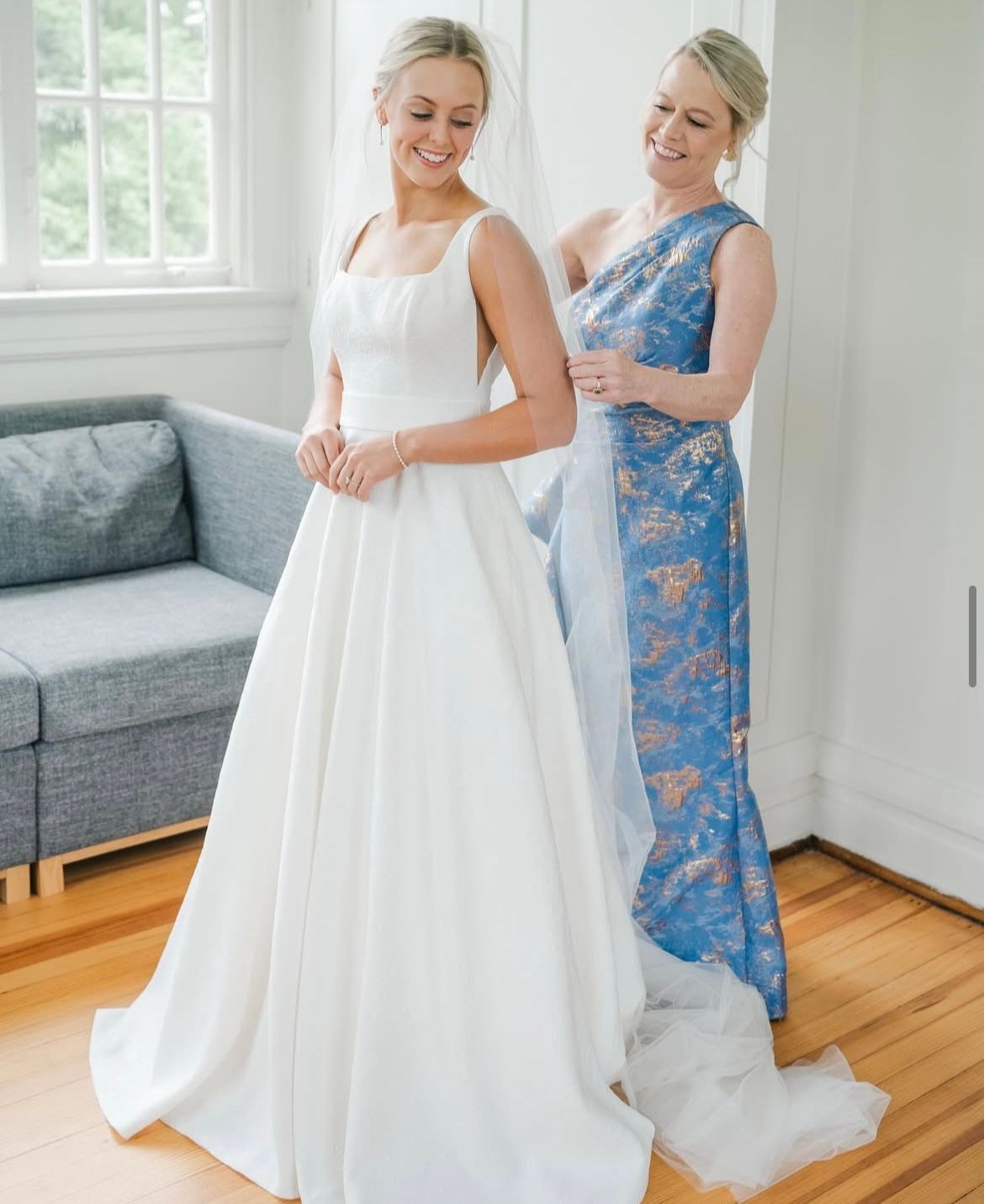 Bride in a white dress, mother helping with veil, blue and gold dress. Inside a bright room with a window.