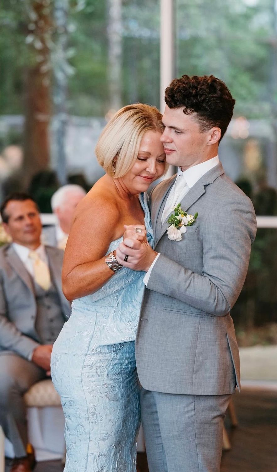 Mother and son dancing, she in light blue dress, he in gray suit, inside, smiling.