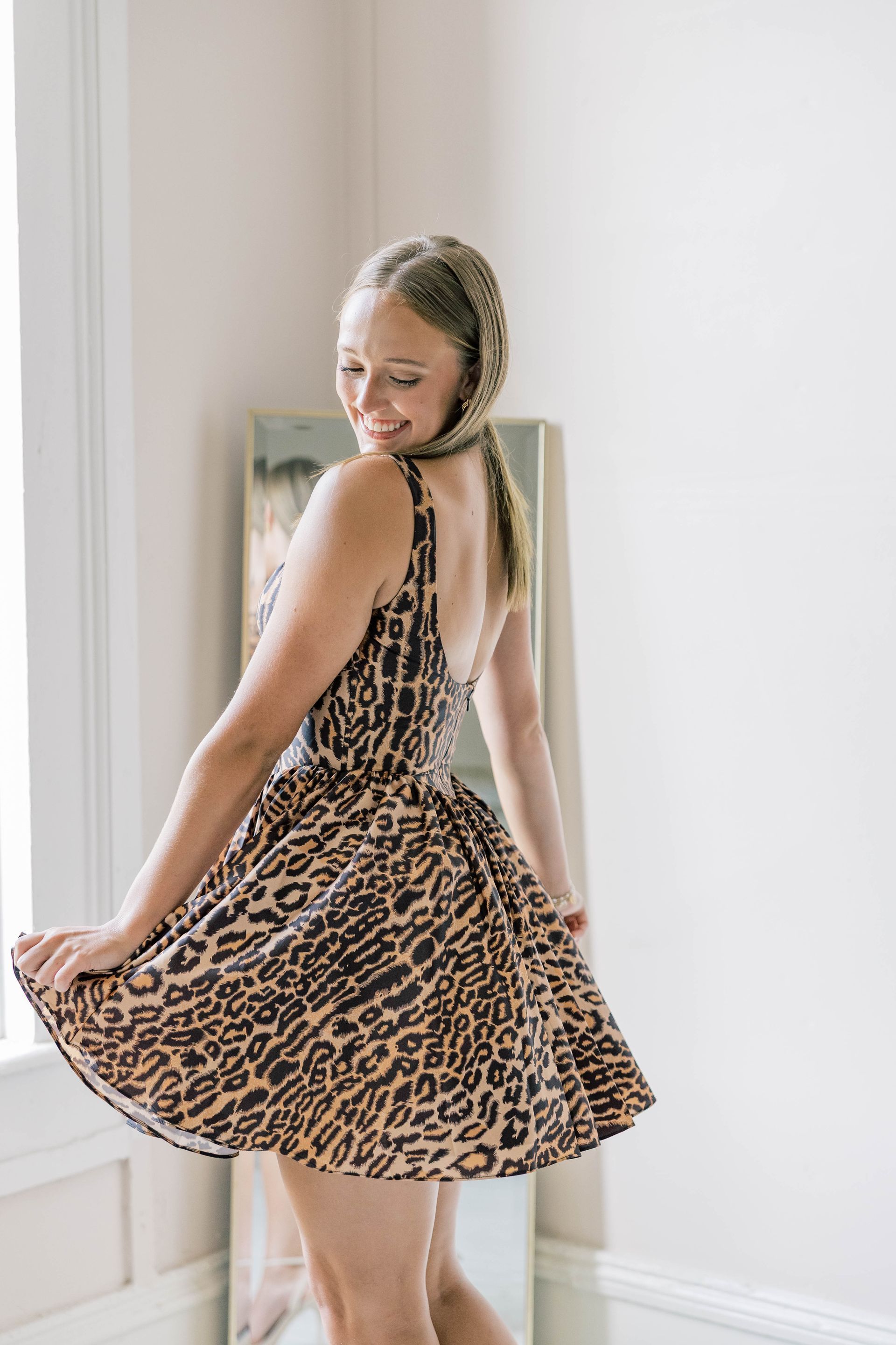 Woman in leopard print dress twirling, smiling at a mirror in a bright room.