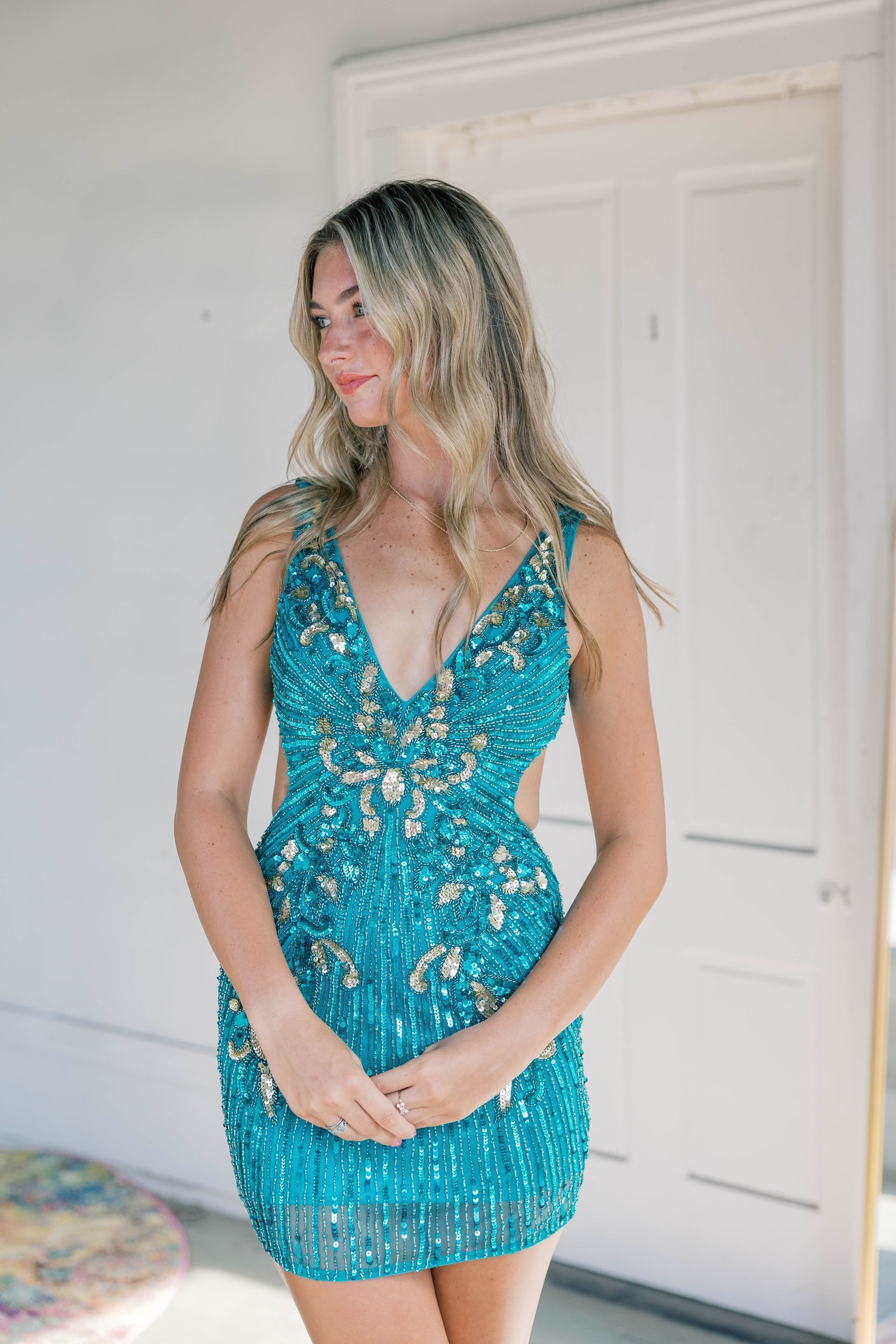 Blonde woman in a turquoise beaded cocktail dress, standing in front of a white door.