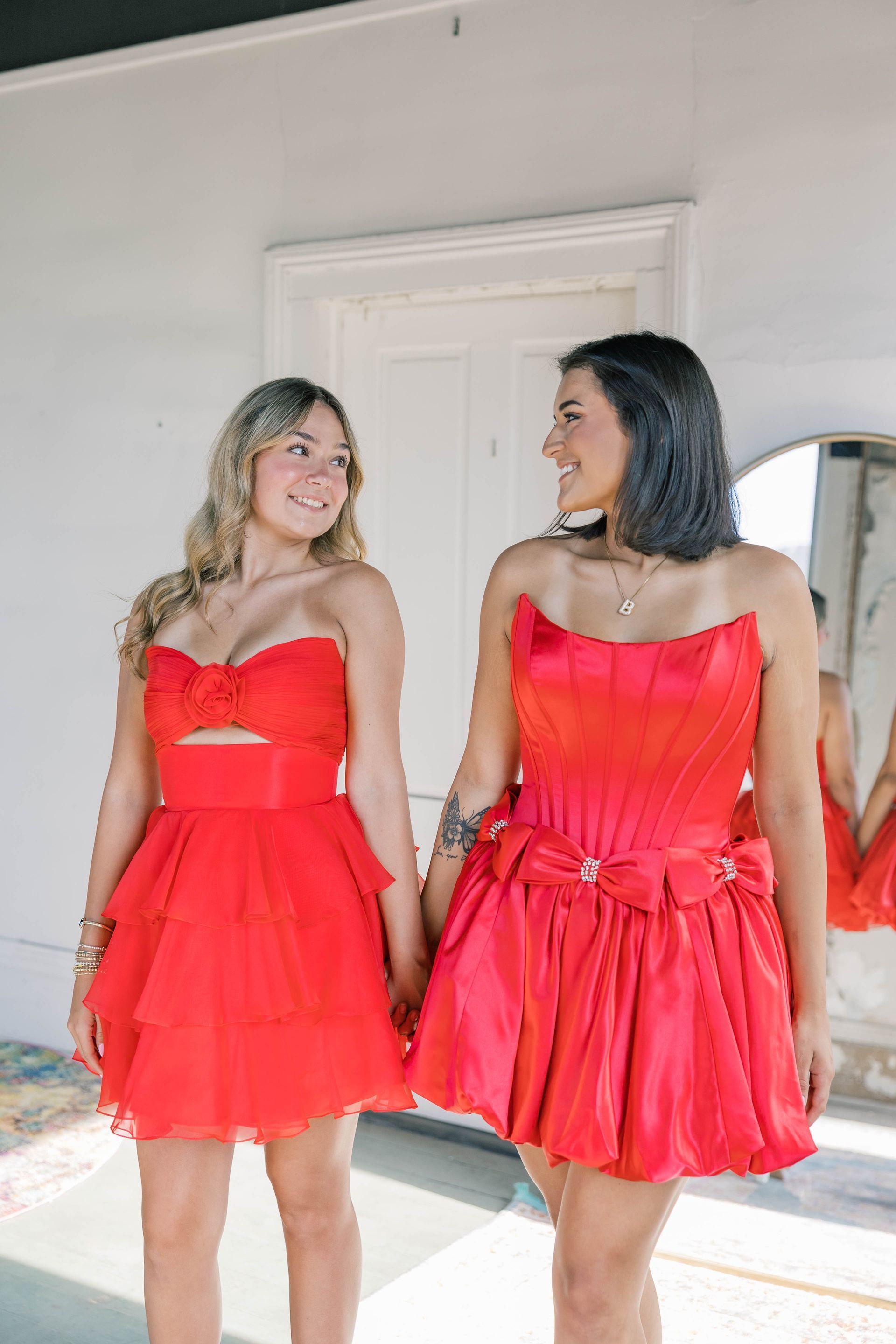 Two women in red dresses holding hands, smiling. Bright room with a door and mirror.