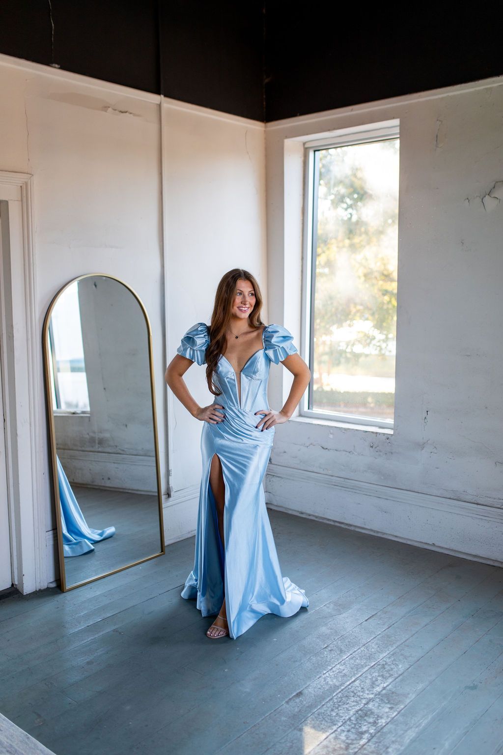 A woman in a light blue dress is standing in front of a mirror in a room.