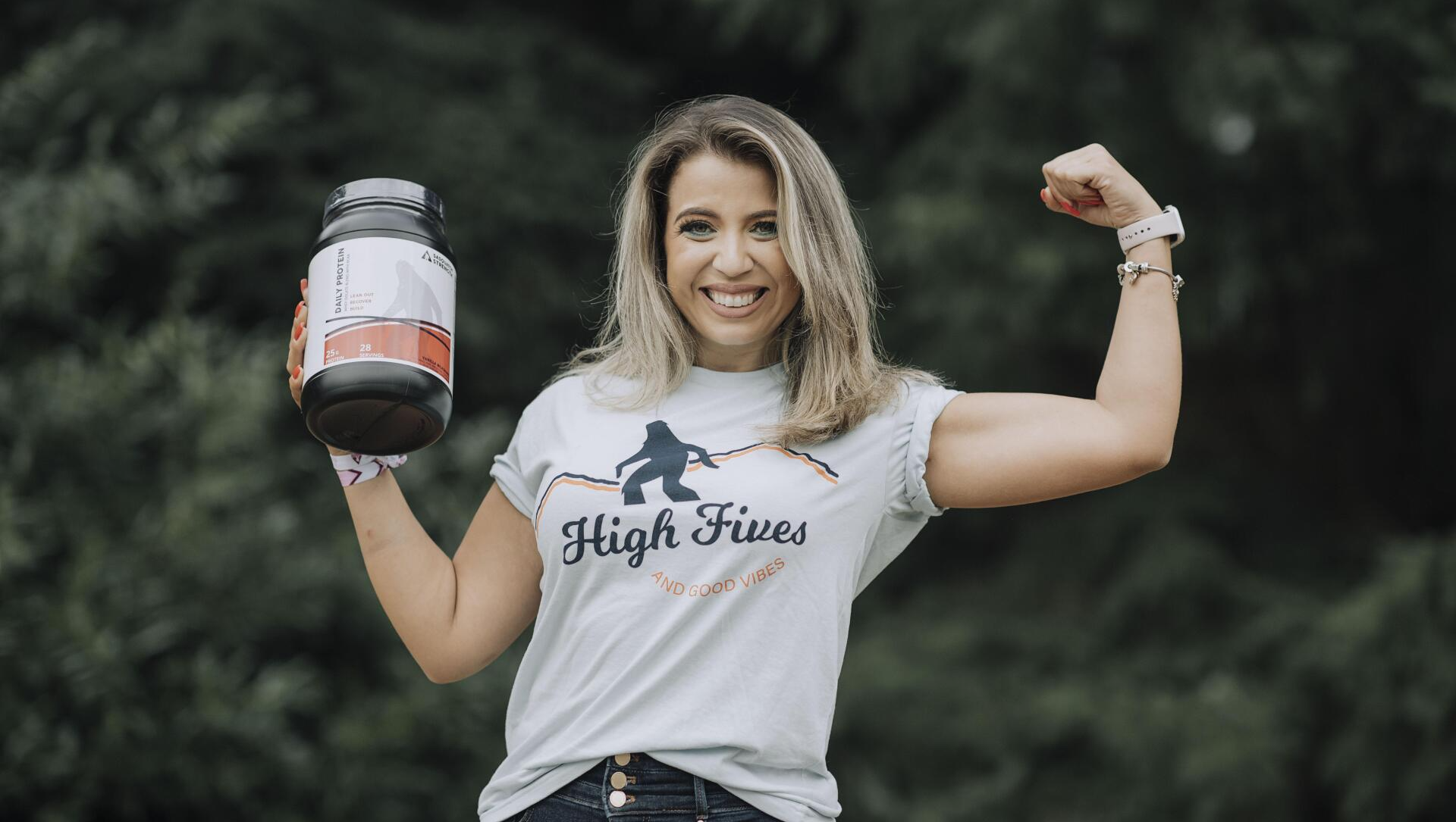 A woman in a high fives shirt is holding a jar of protein powder.