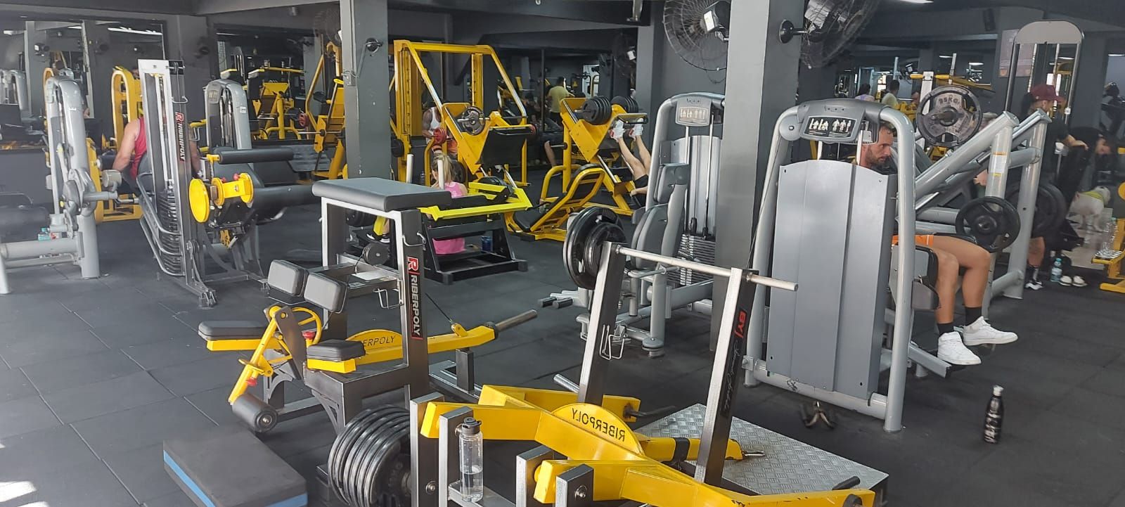 Interior de academia com vários aparelhos de musculação amarelos e cinzas. Uma pessoa usa um aparelho de supino sentado.