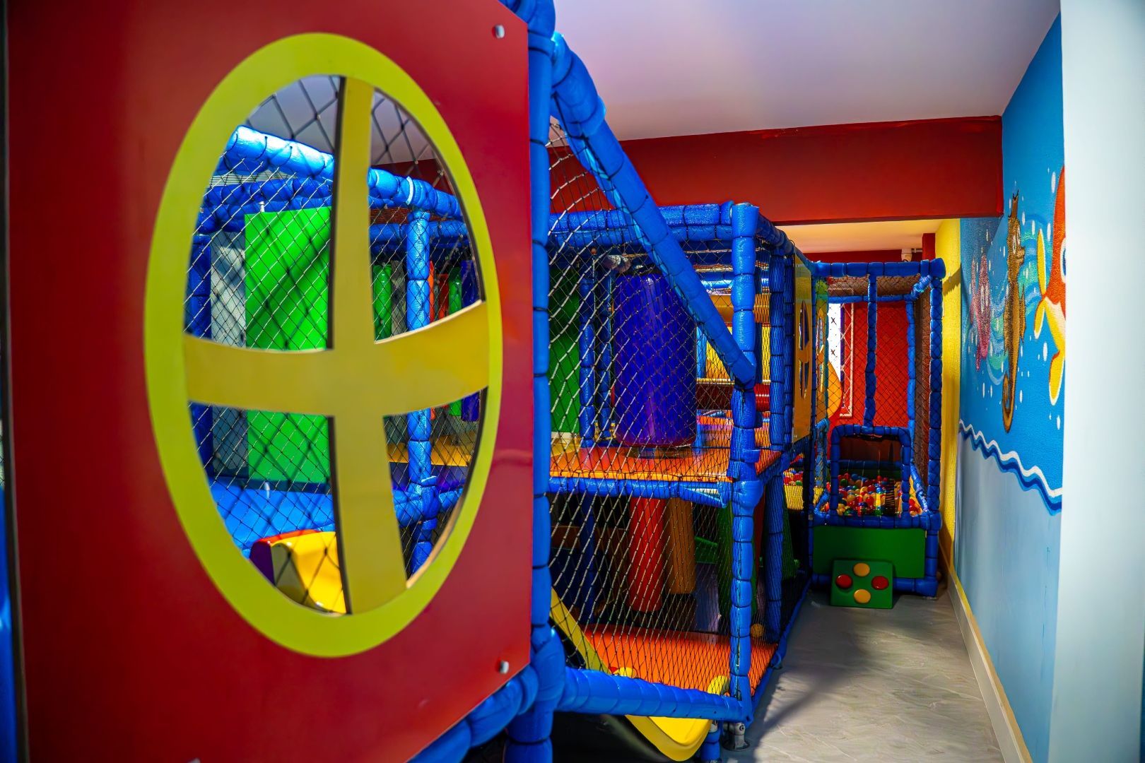 Área de recreação infantil interna com estruturas de escalada coloridas, redes e uma grande janela.