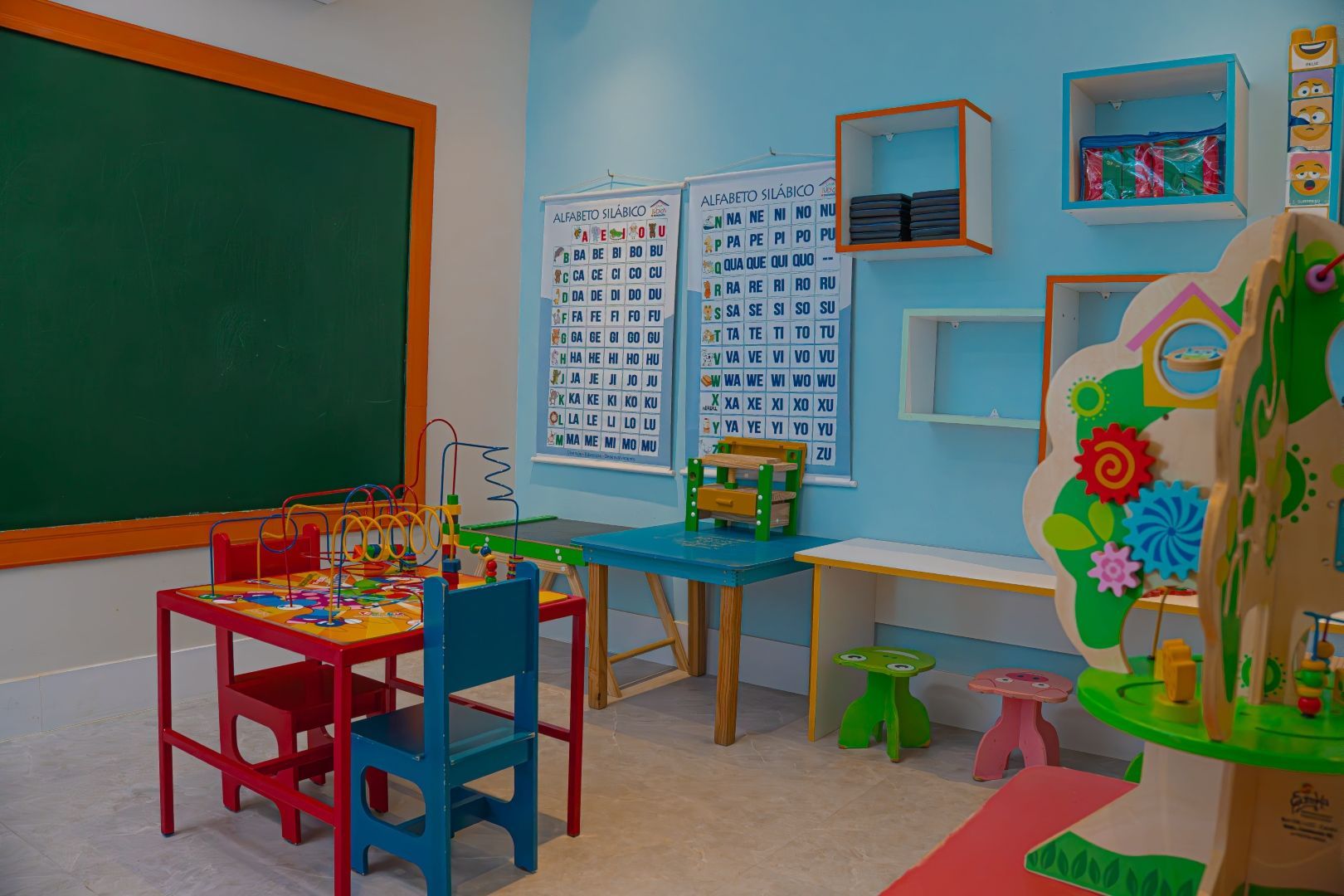 Uma sala de jogos infantil colorida com quadro-negro, mesas, cadeiras, alfabeto e prateleiras.