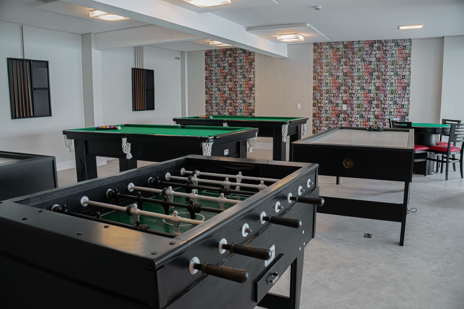 Uma sala de jogos com pebolim, mesas de sinuca e shuffleboard. Possui paredes com padrões coloridos e paredes brancas.