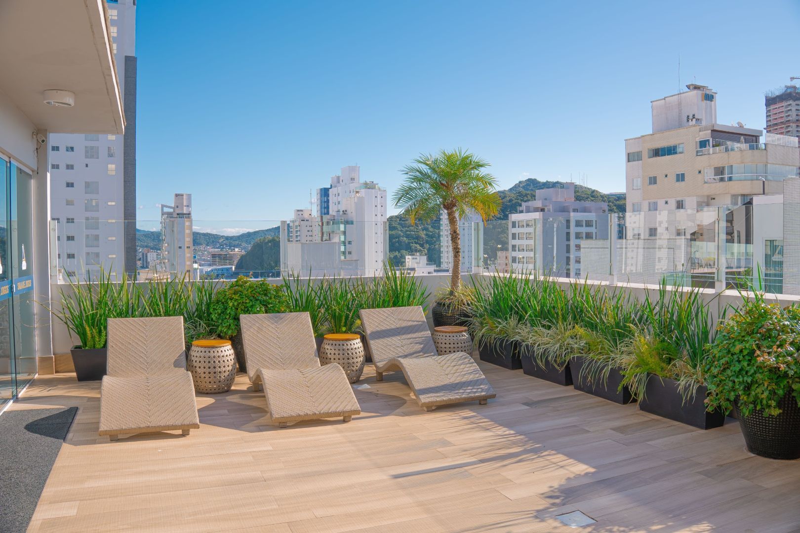 Pátio na cobertura com espreguiçadeiras, plantas e vista para a cidade em um dia ensolarado.