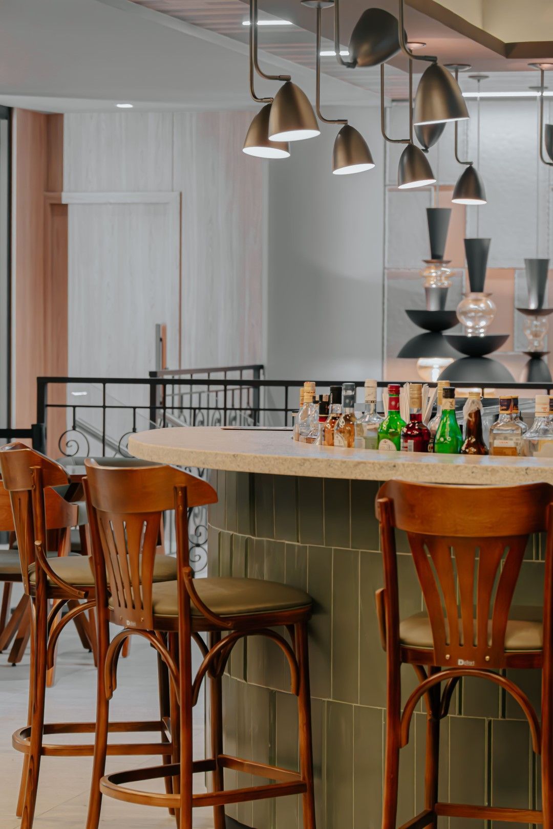 Bar com bancos de madeira, garrafas e iluminação decorativa. Azulejo verde na base do bar, paleta de cores neutras.