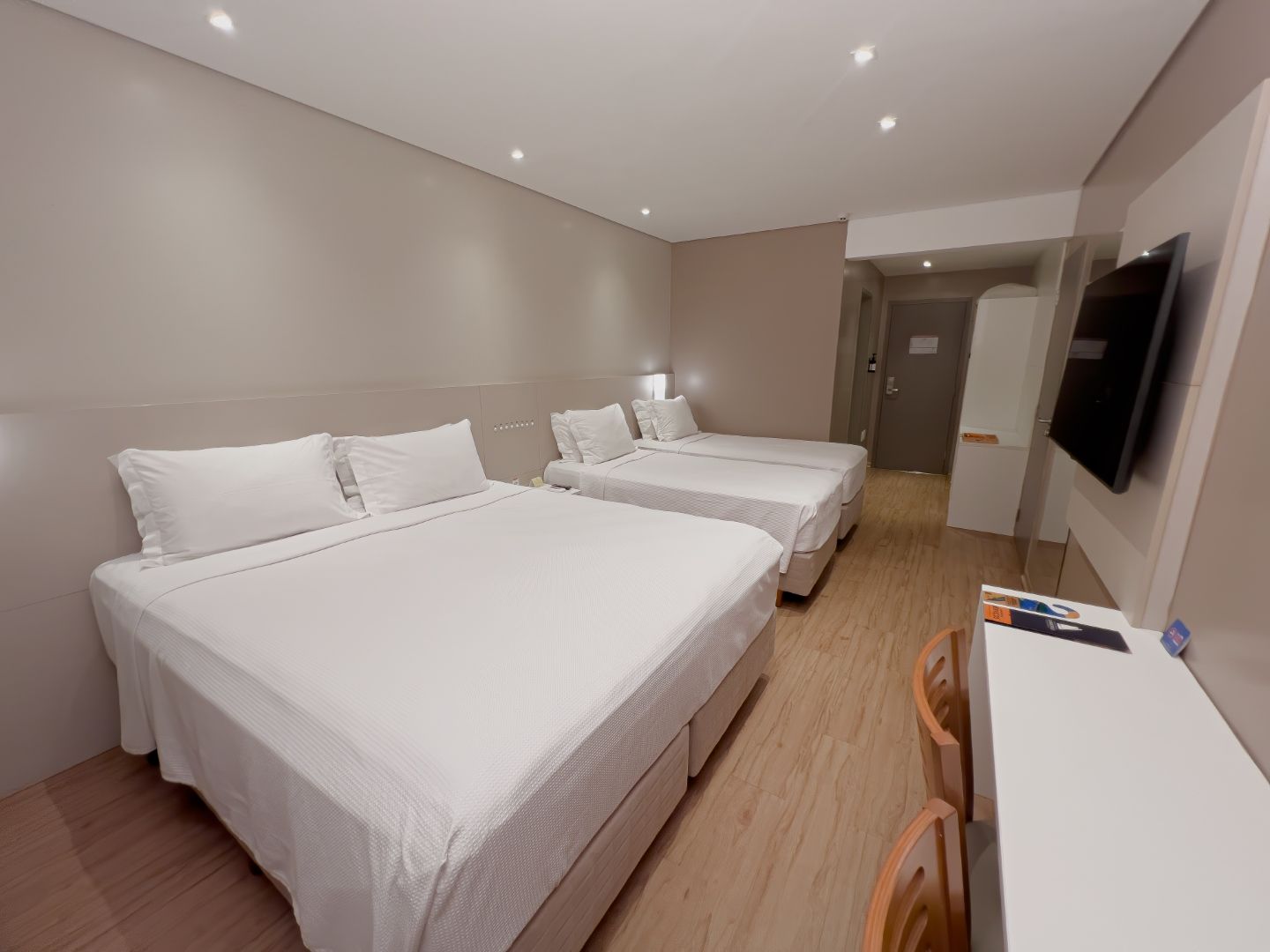 Quarto de hotel com três camas, paredes neutras, TV e uma escrivaninha.