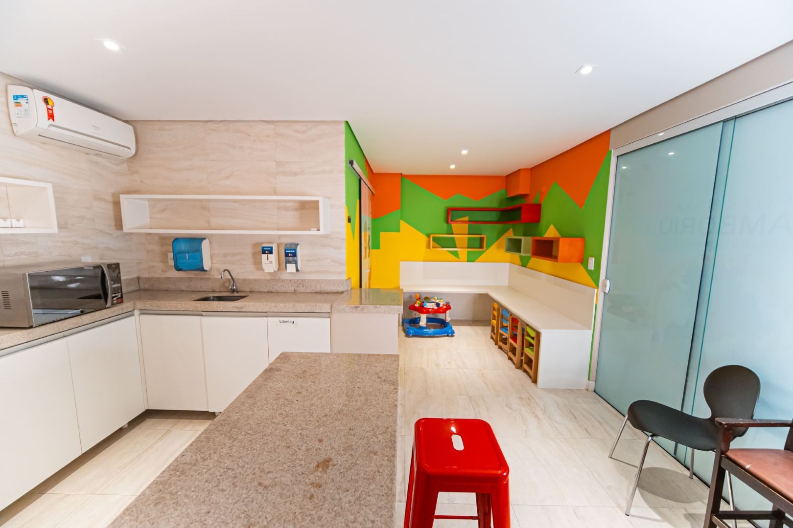 Área de cozinha e sala de jogos: armários brancos, bancada, micro-ondas, paredes coloridas, banco vermelho, área de jogos visível.